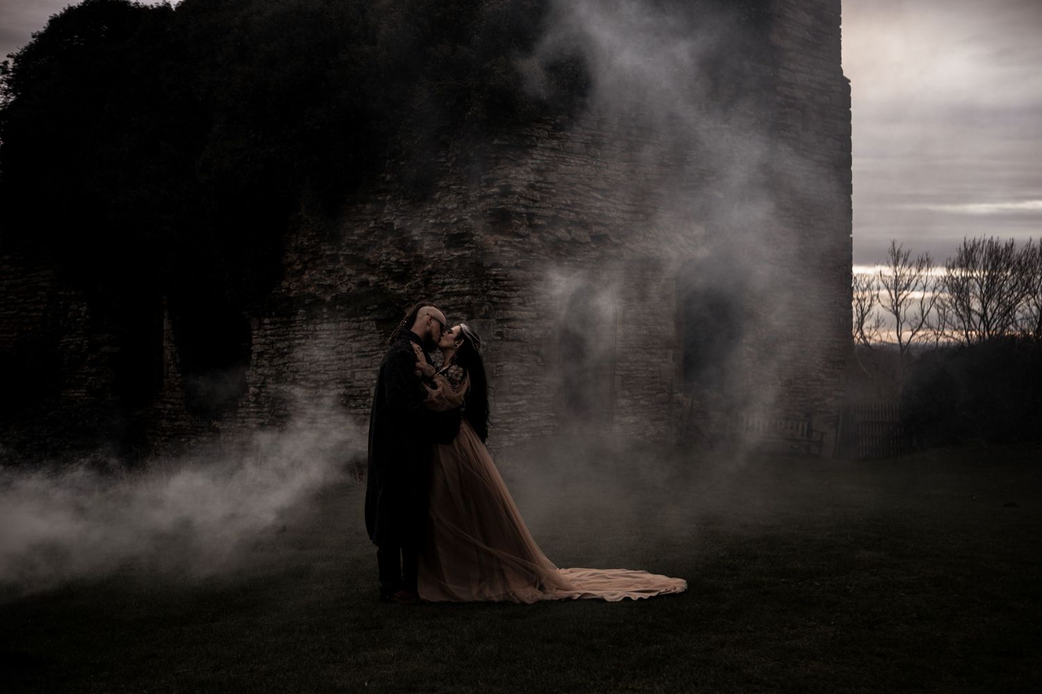 Gothic wedding couple embracing by ancient stone ruins surrounded by atmospheric mist at dusk.