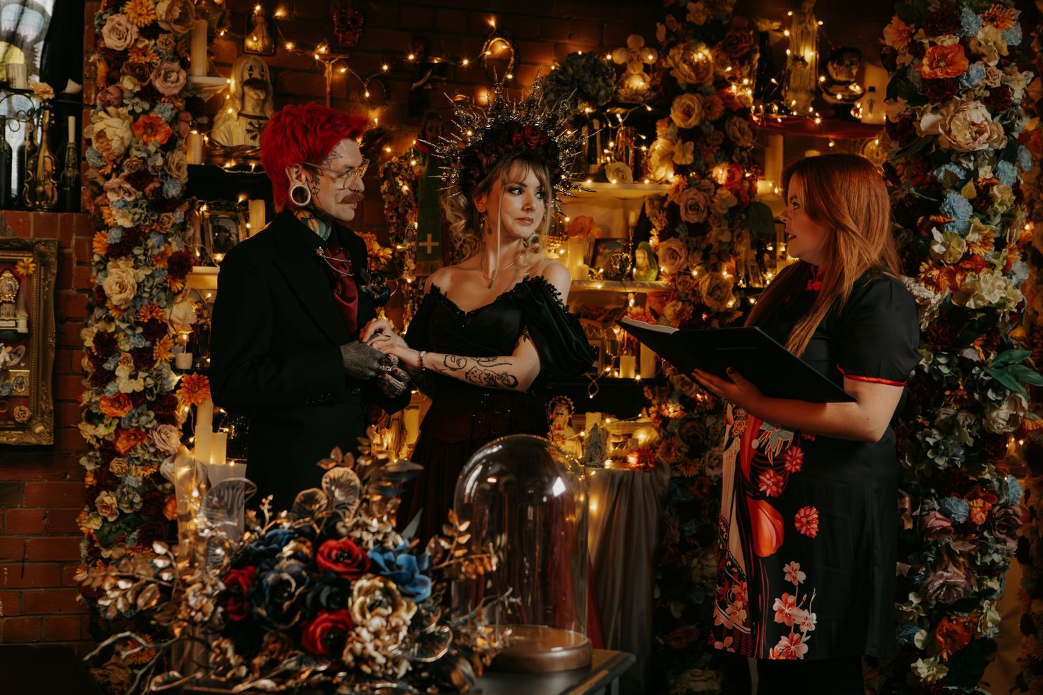 Couple exchanging vows at colorful floral altar with officiant in warmly lit bohemian venue.