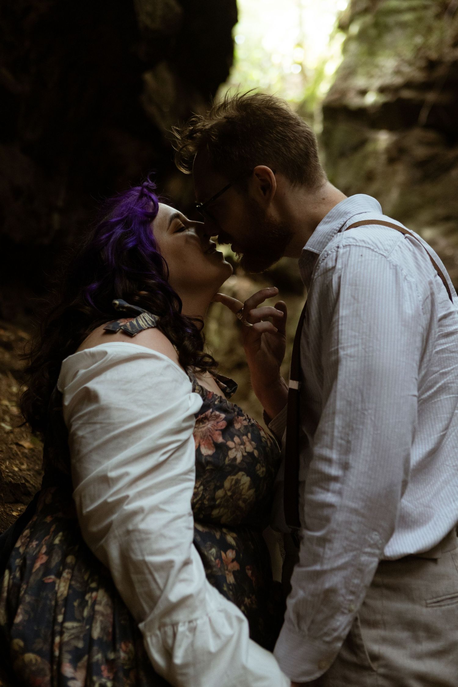 A couple with purple hair and suspenders share a romantic moment in a moody forest cave setting.