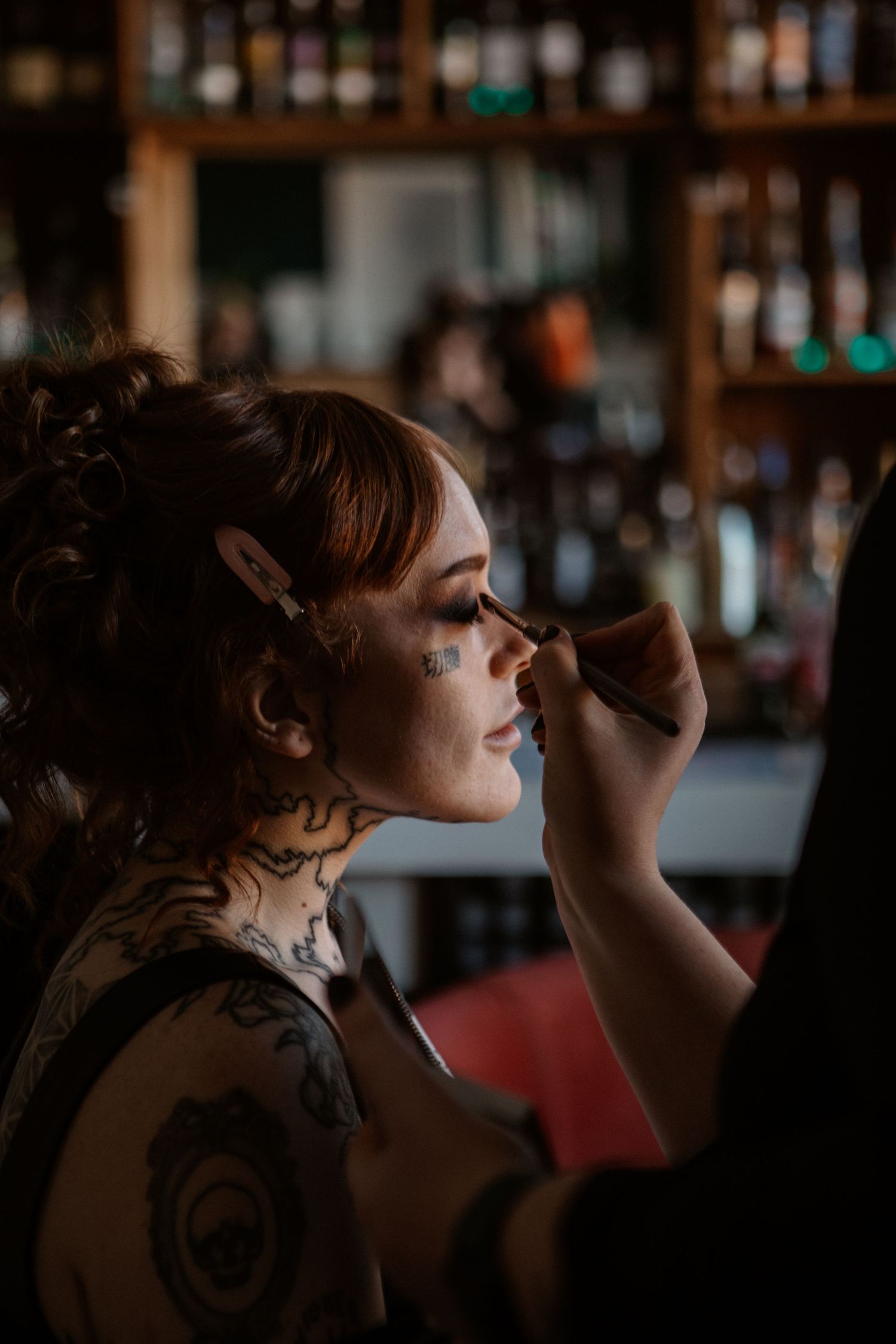 Woman with neck tattoos and face gems getting eye makeup applied by a makeup artist in a bar setting.