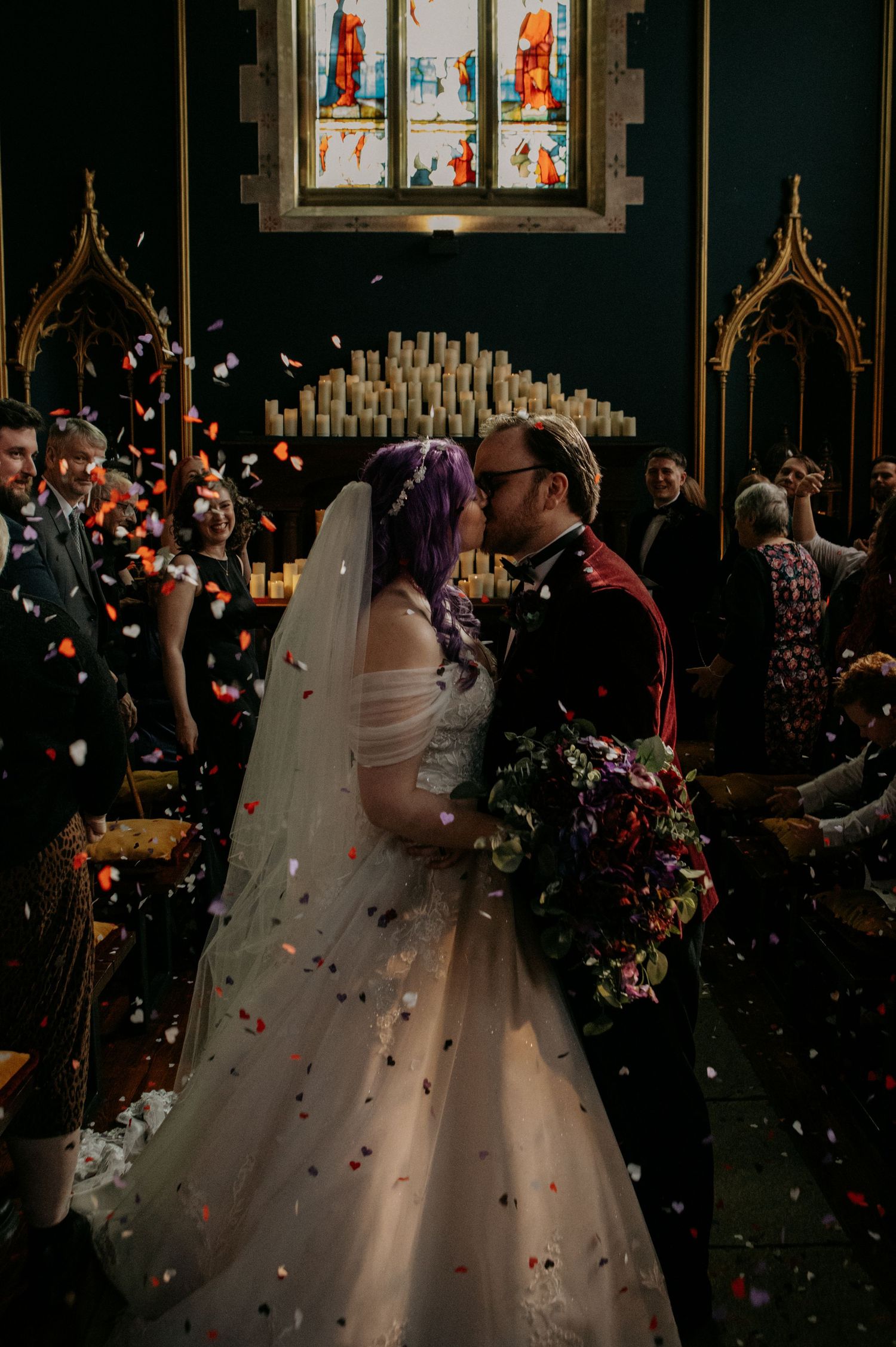 Bride with purple hair and groom share first kiss at church altar as colorful confetti fills the air.