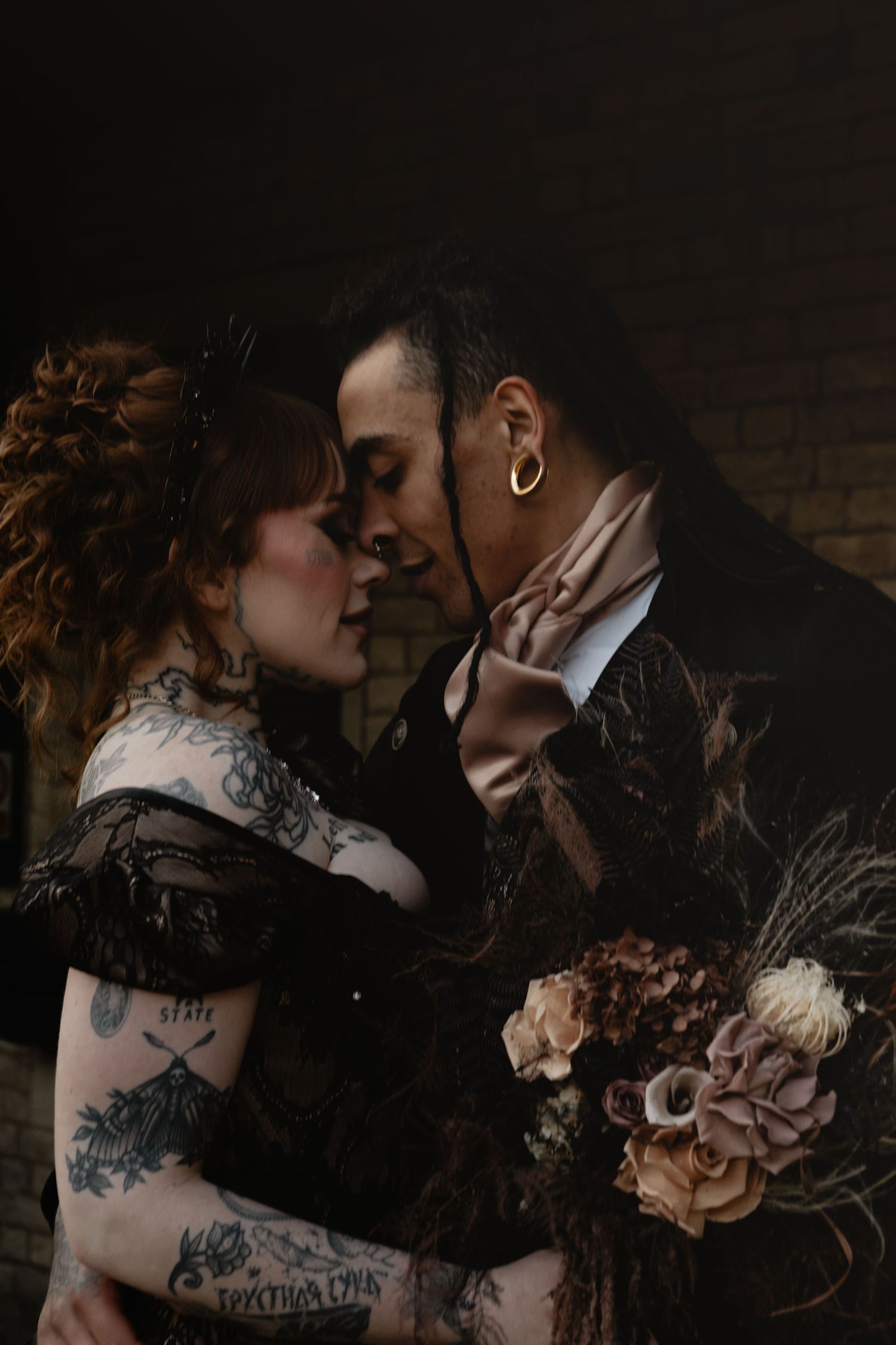 A tattooed couple in gothic wedding attire share an intimate moment, holding a dark floral bouquet against a moody backdrop.
