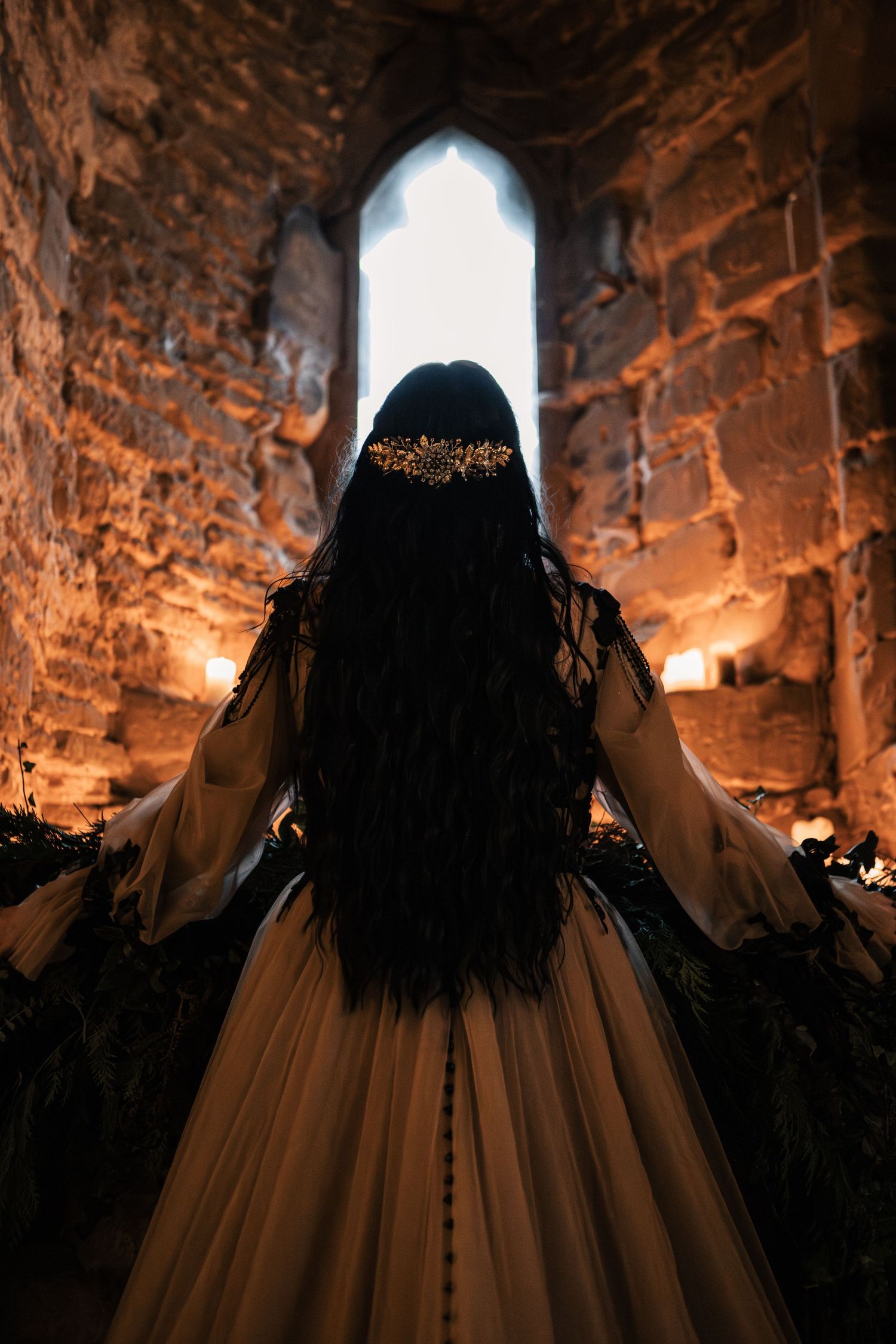 Gothic bride in golden gown with black feathers facing arched window in candlelit stone ruins.