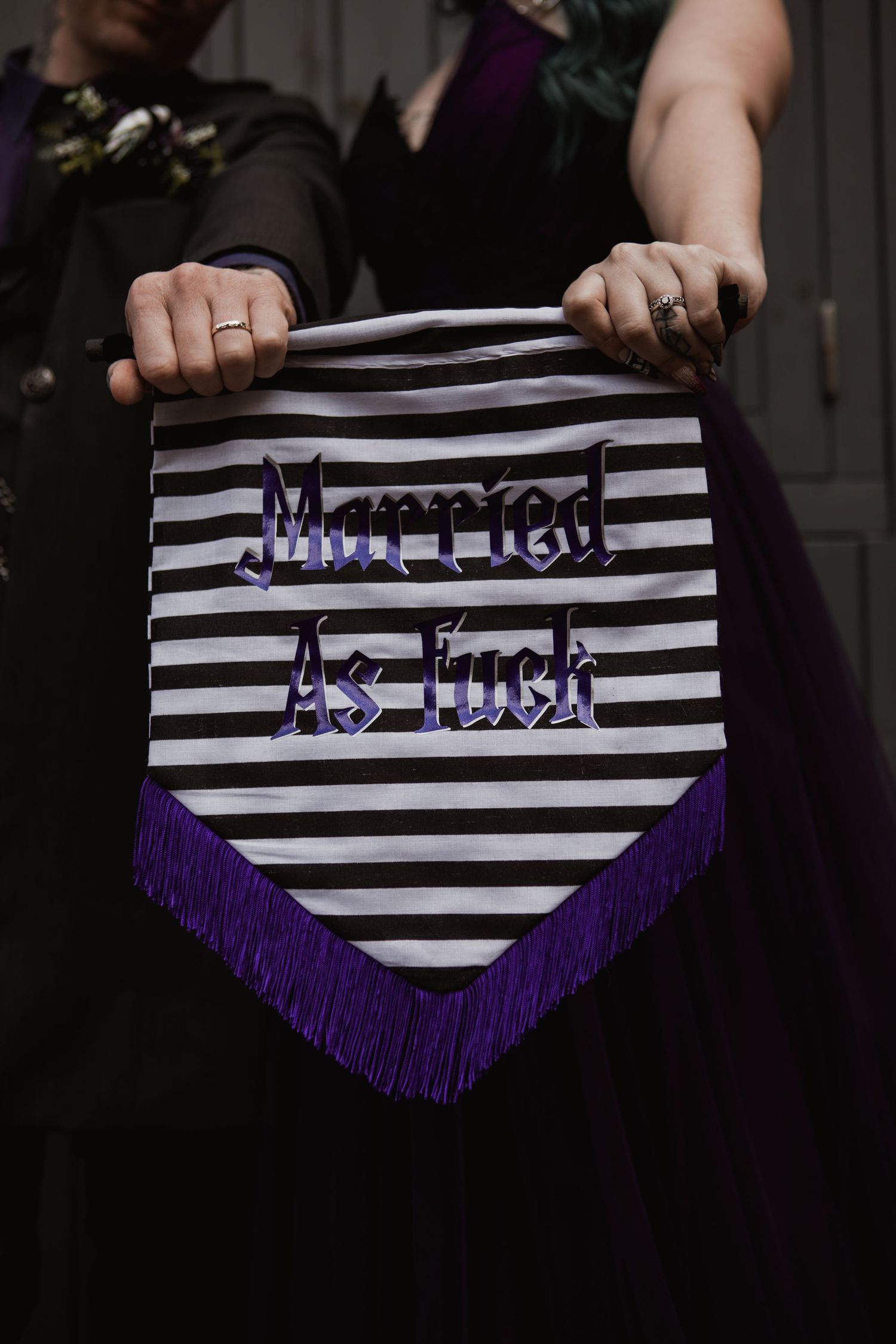 Black and white striped pennant banner with purple fringe reading Married As Fuck held by newlyweds.