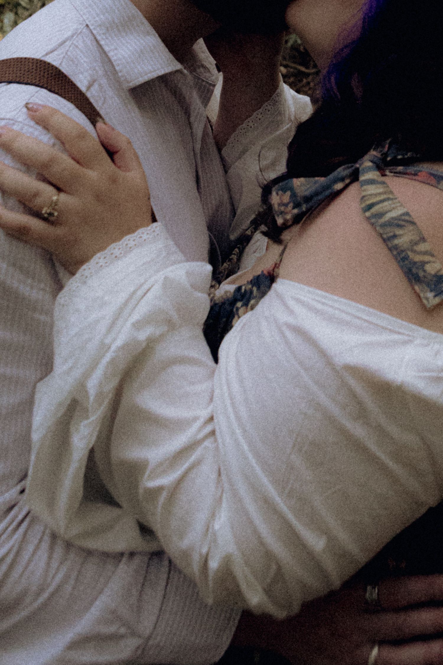 A couple embraces tenderly, the woman wearing a white off-shoulder dress with a tattoo visible on her shoulder.