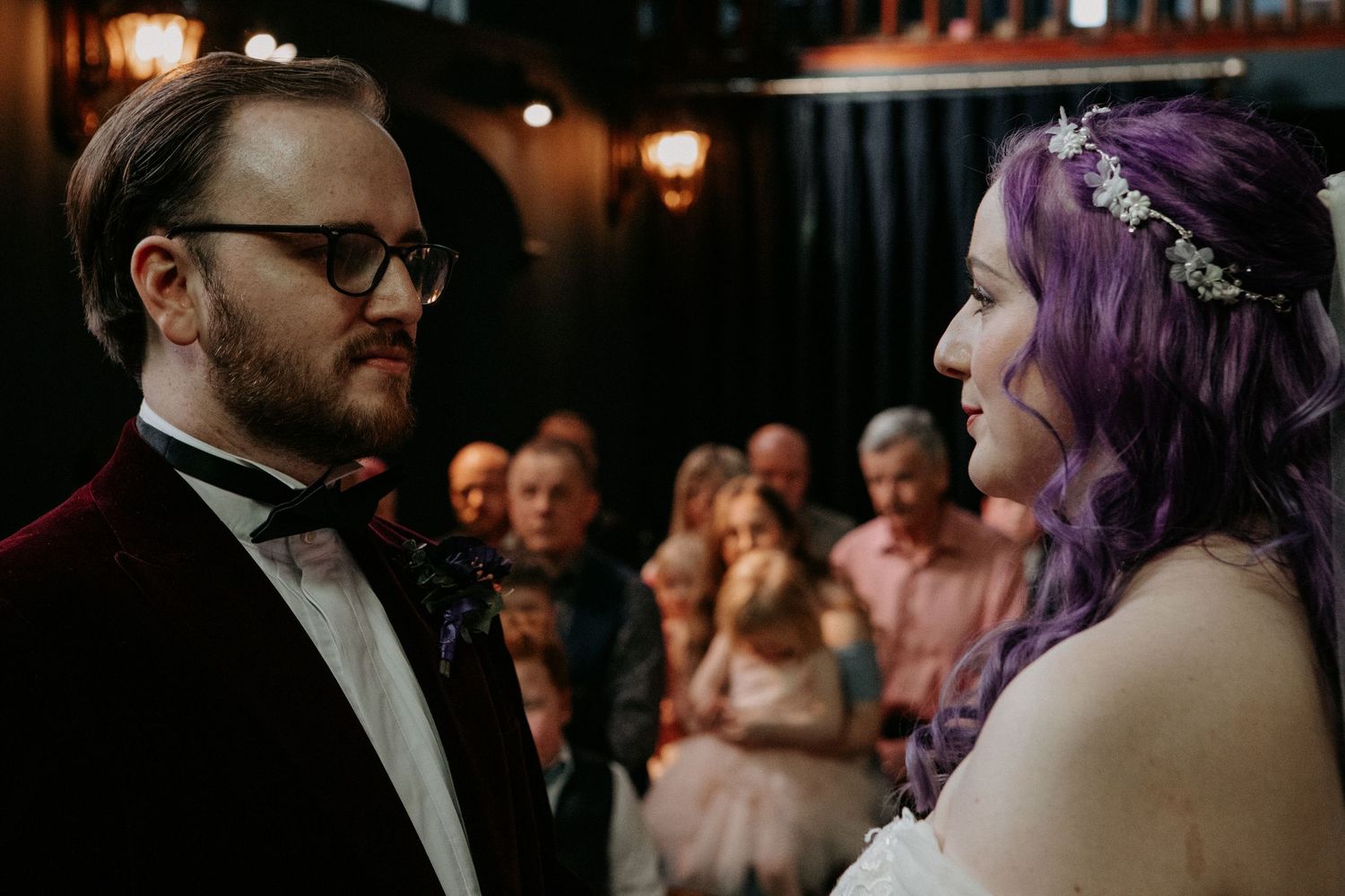 Couple exchanging vows at wedding ceremony, groom in tuxedo, bride with purple hair and floral crown, guests in background.
