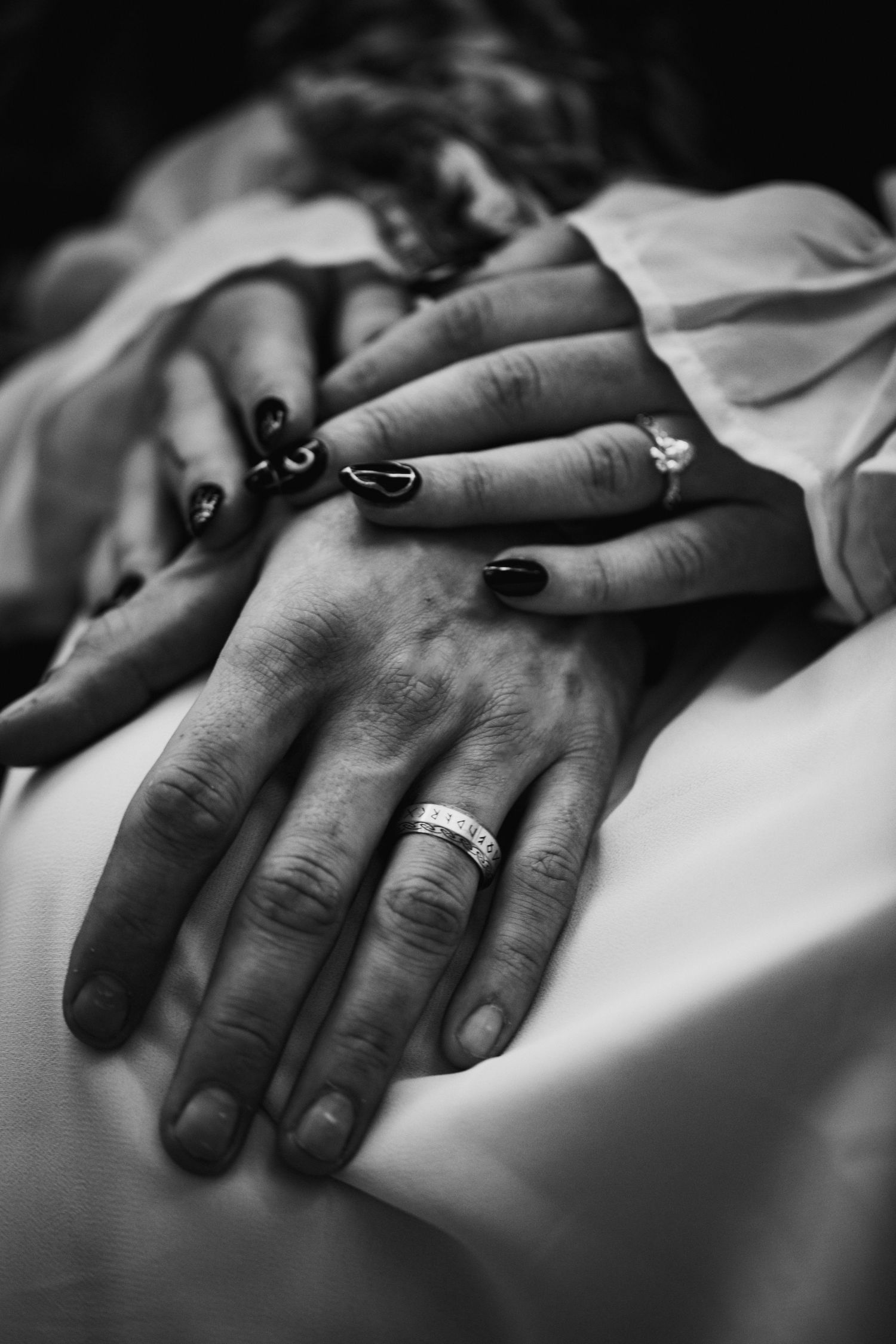 Black and white close-up of hands with dark nail polish and a wedding ring resting on white fabric.