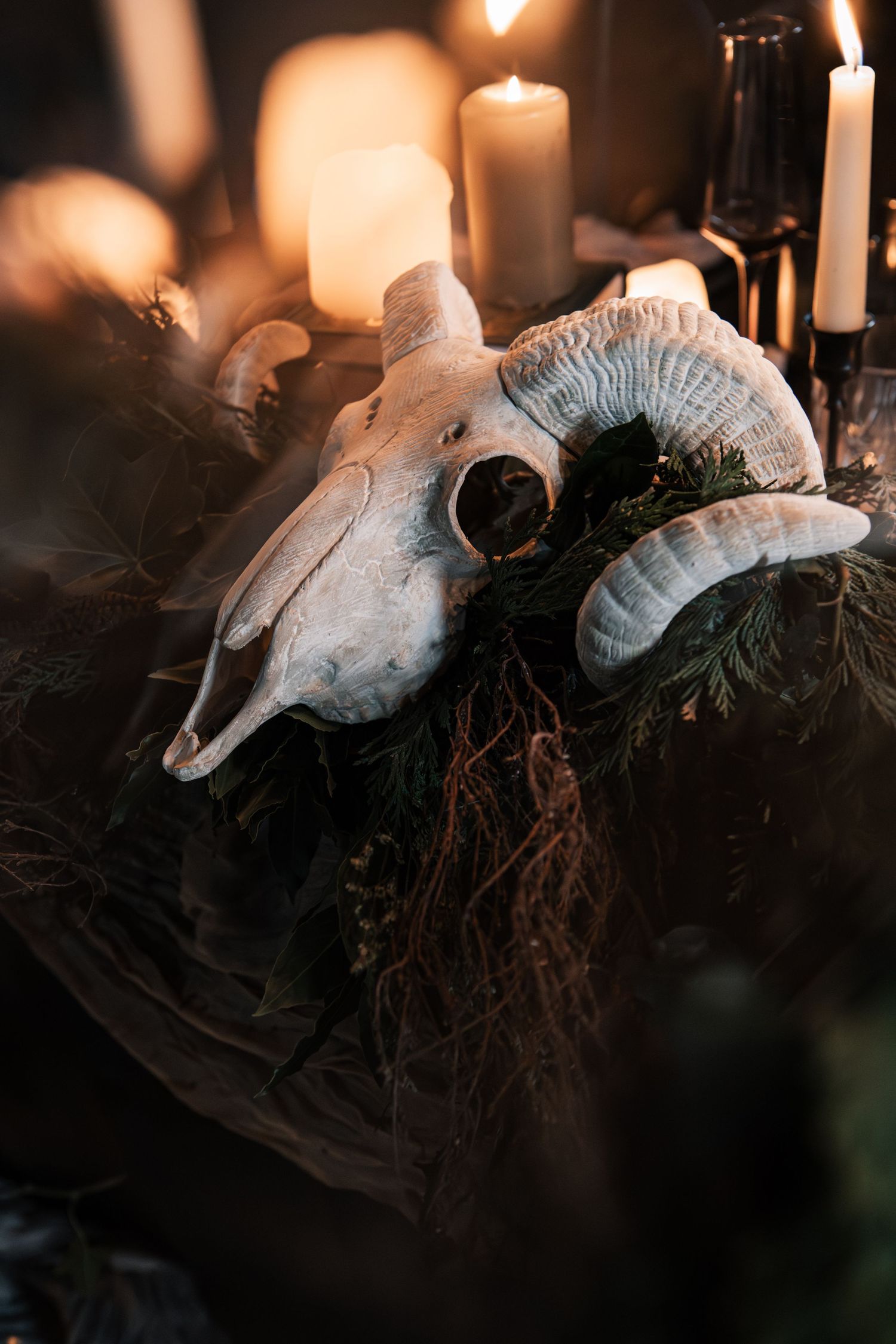 A ram skull with curved horns surrounded by greenery and glowing candles in a dark, moody occult display.