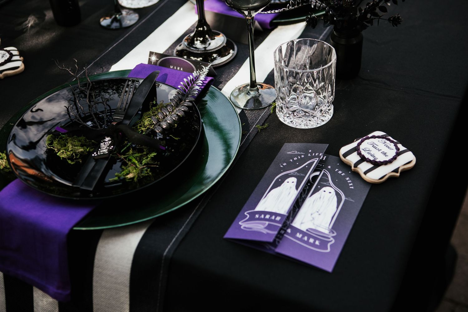 Elegant Halloween table setting with black and purple accents, crystal glassware, dark floral decor, and spooky themed menu cards.