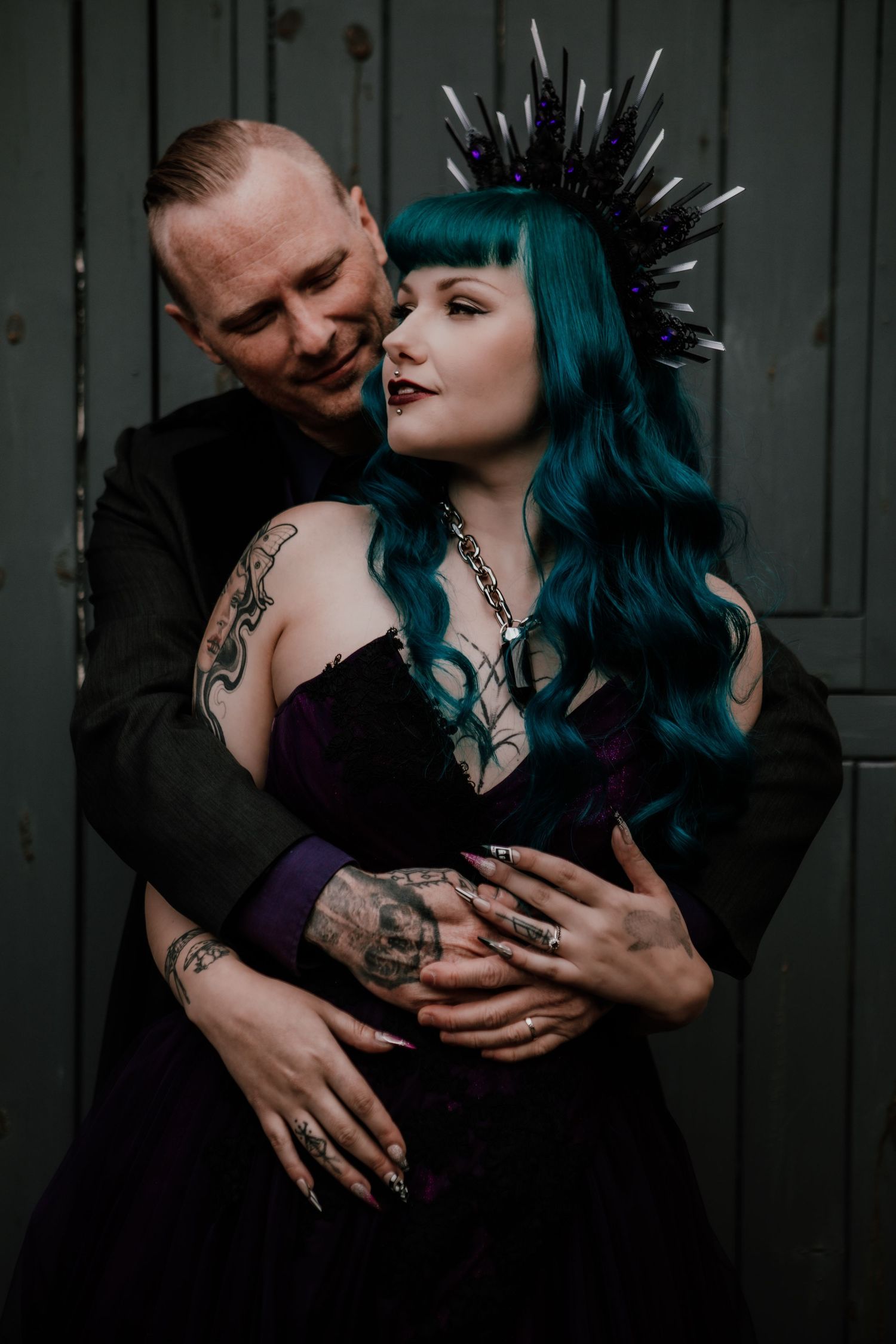 Gothic couple embracing, woman with teal hair and spiked crown in purple corset, tattooed man in black suit, dark moody backdrop.
