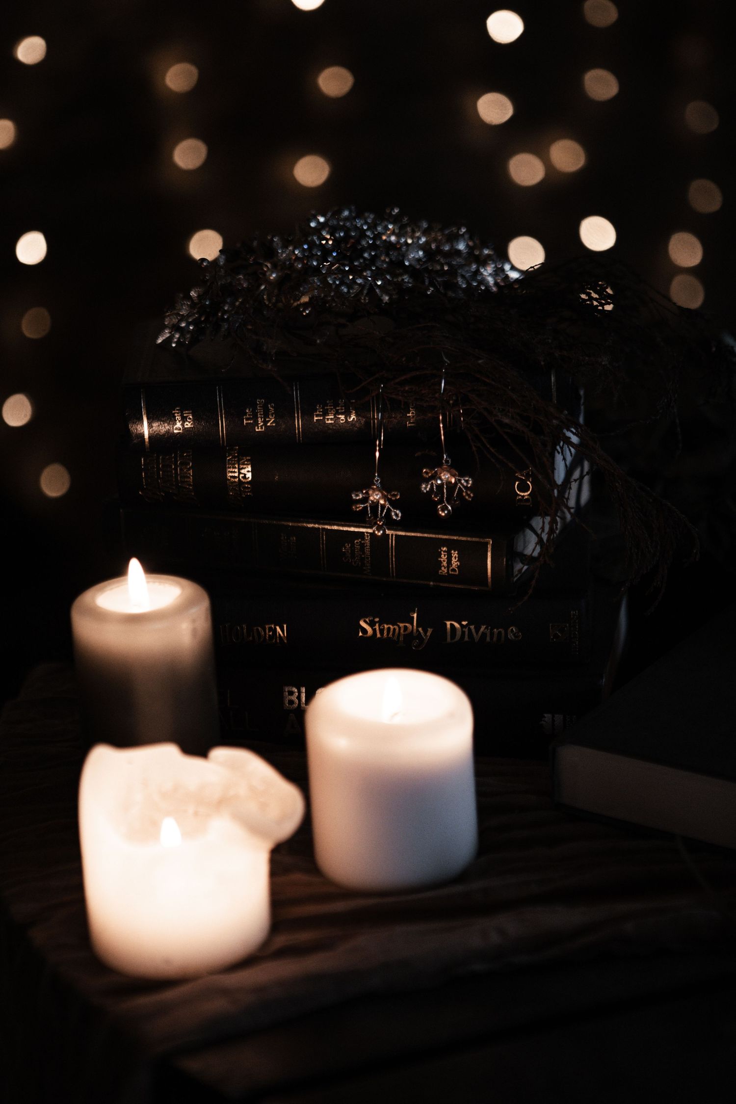 Dark gothic scene with glowing candles, black books titled Simply Divine, and bokeh lights in background.
