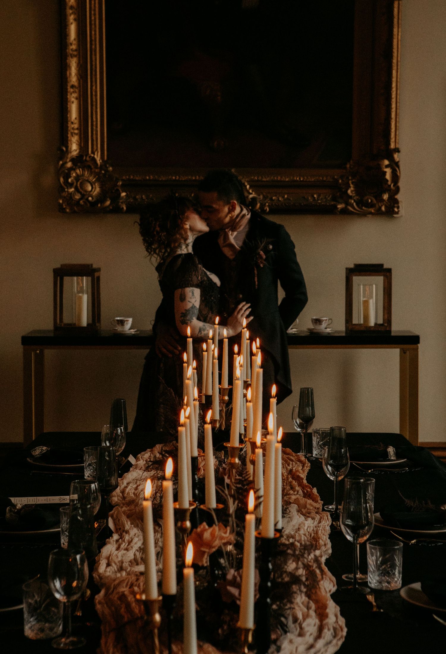 Gothic wedding couple kissing behind tall white taper candles at dark romantic candlelit reception table.