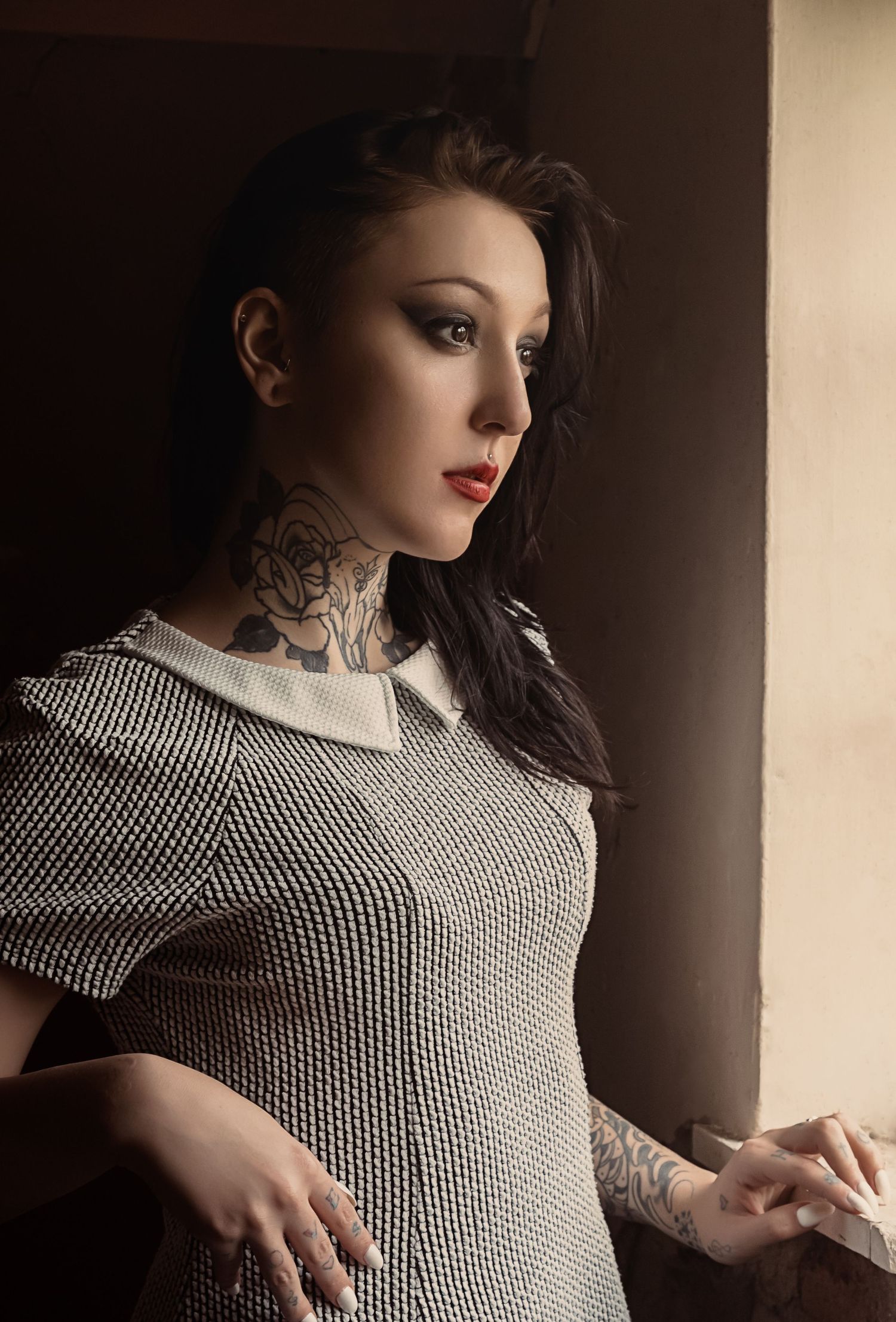 Romantic portrait in dramatic lighting shows a figure in gingham dress with neck tattoos gazing thoughtfully by a window.