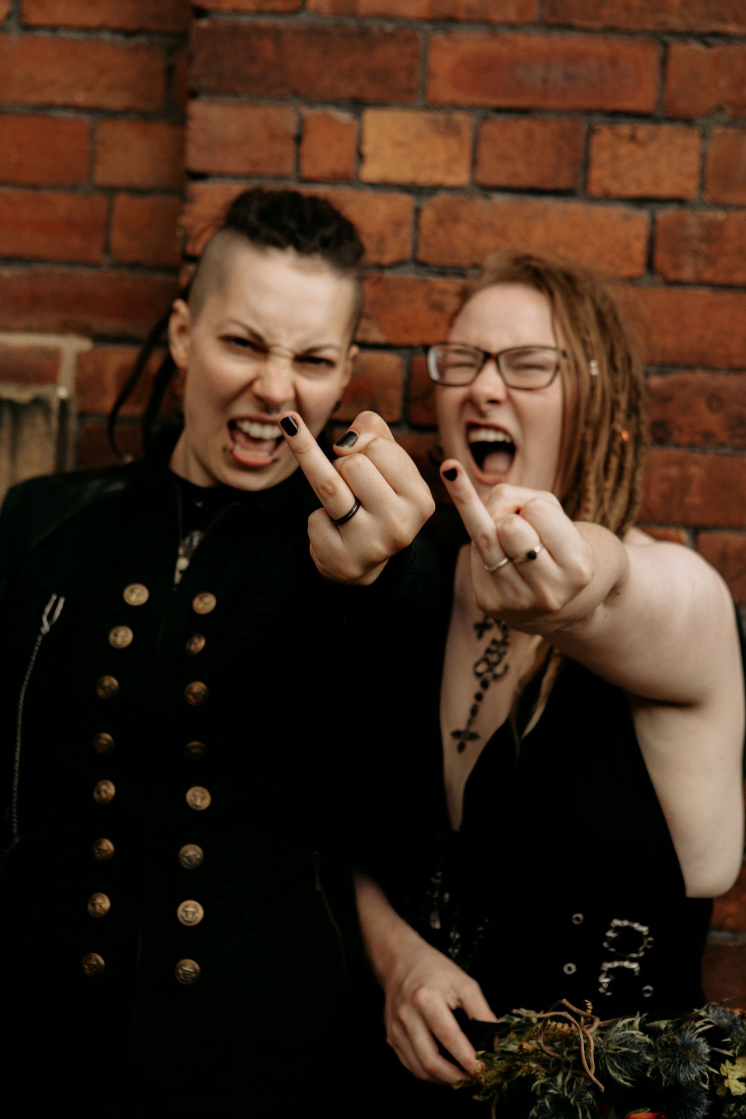 Two women in black gothic attire making rock hand gestures in front of a brick wall.