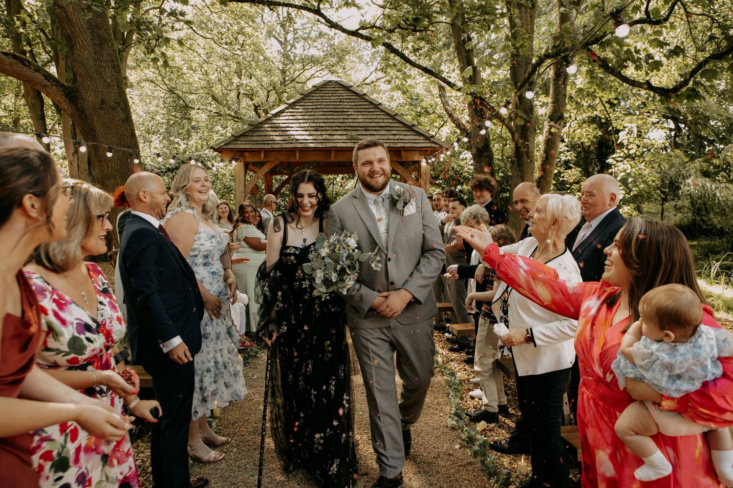 Bride in black gown and groom in gray suit walk through confetti shower among guests at outdoor woodland wedding ceremony.