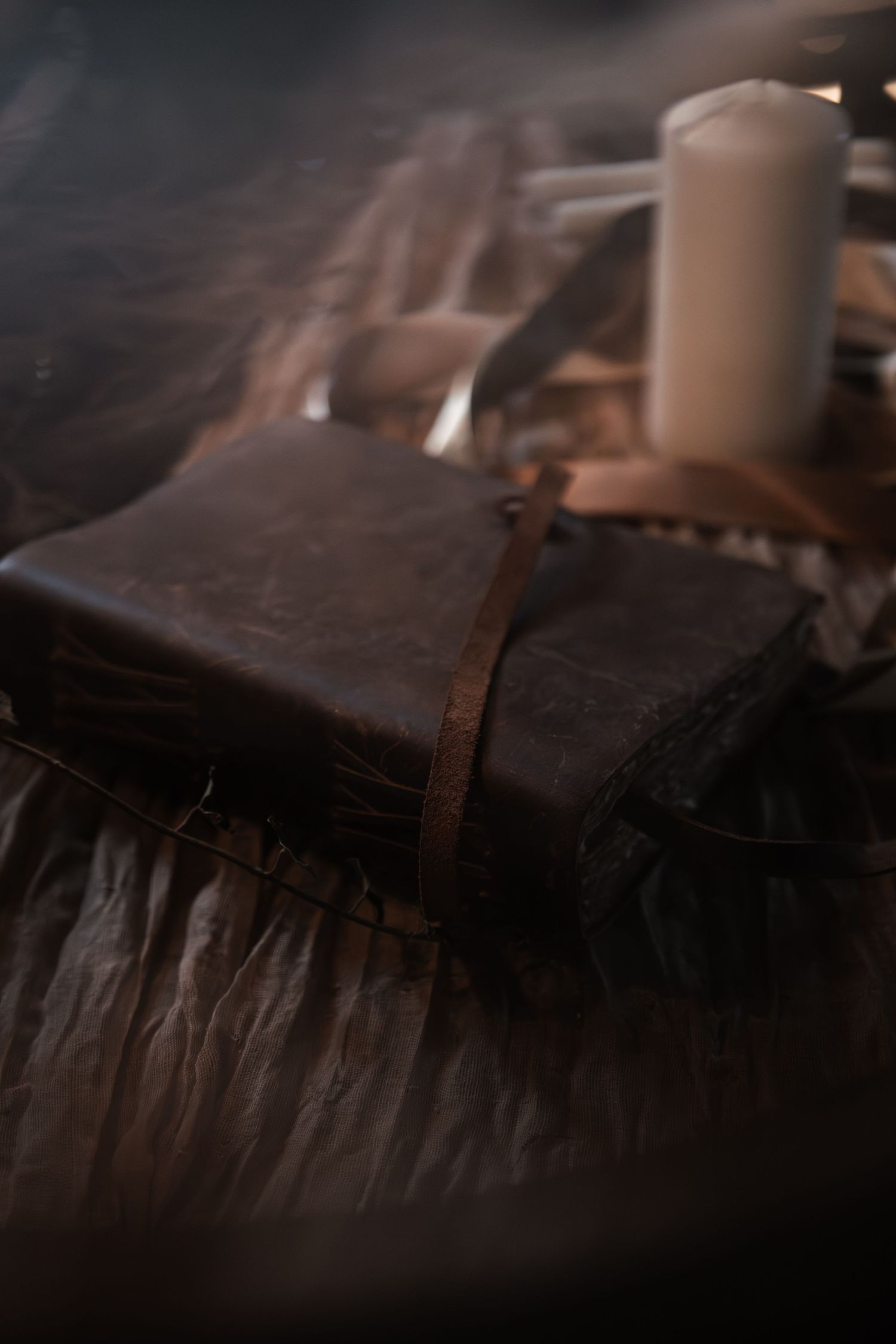 Aged leather-bound journal with strap closure beside a white pillar candle in a dark, moody setting.