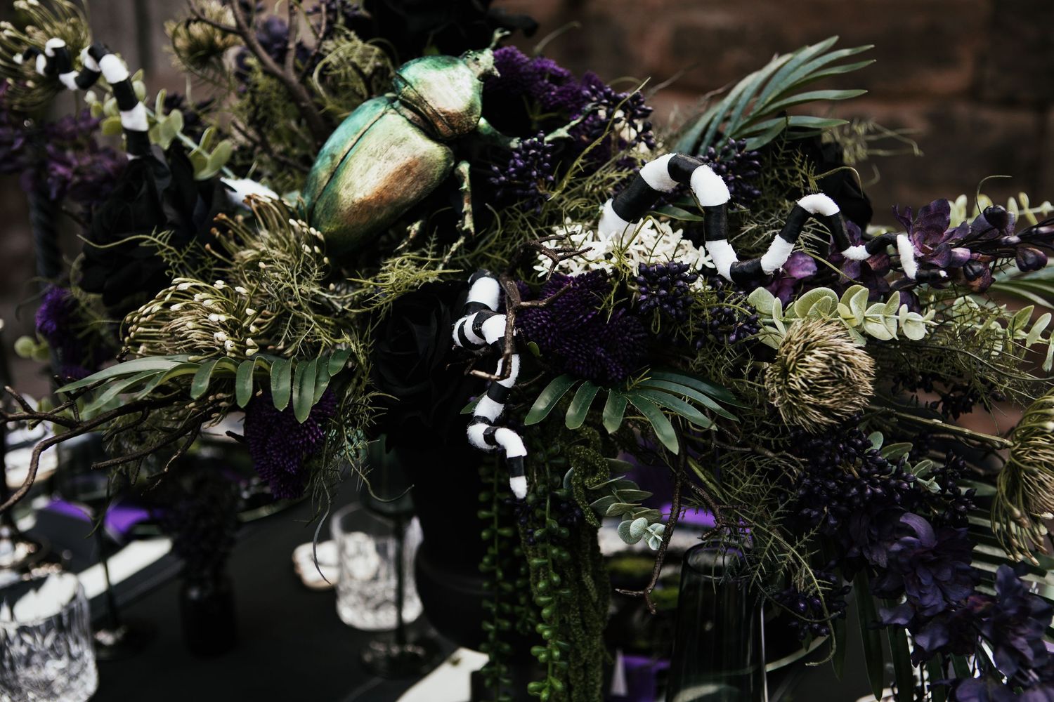 Dark gothic floral arrangement featuring deep purple blooms, tropical leaves, iridescent pods, and black-and-white striped accents.
