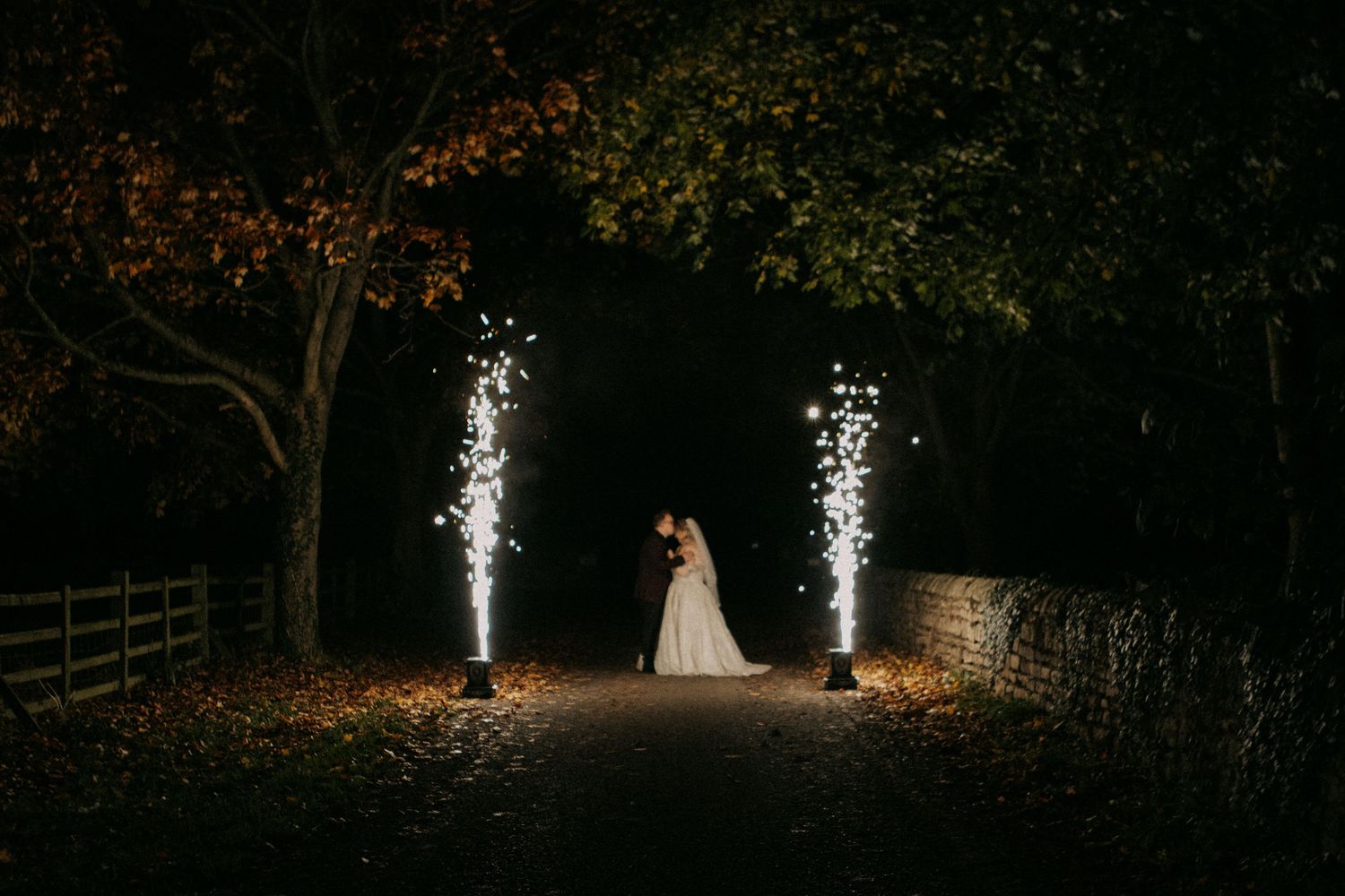 Bride and groom kissing between two sparkling cold fireworks on a tree-lined path at night.