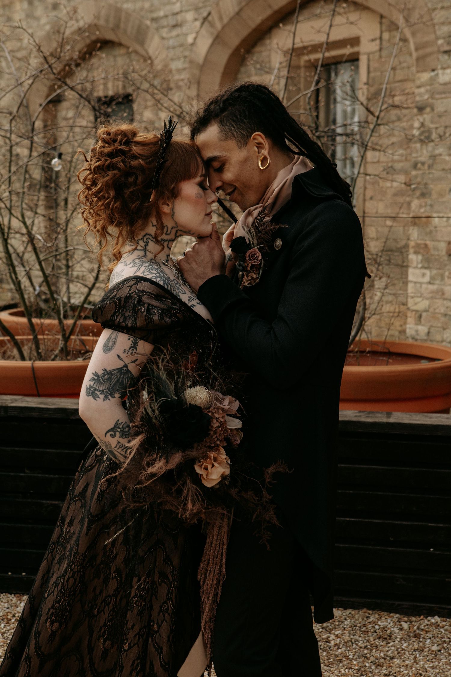 Romantic couple in dark vintage clothing embrace against stone wall backdrop with moody lighting and gothic aesthetic.