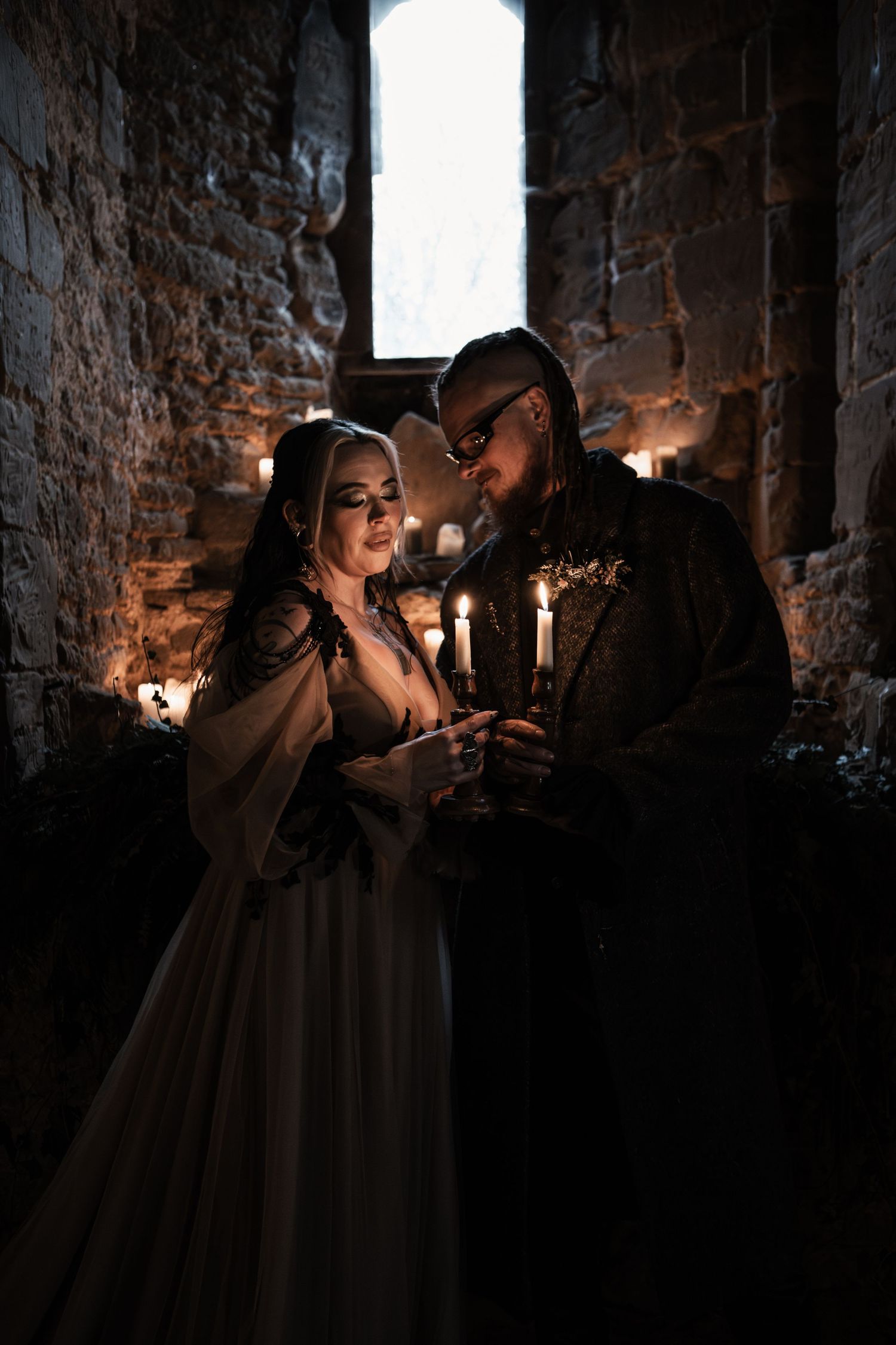 Gothic bride and groom sharing intimate candlelit moment together in dramatic stone castle wedding venue.