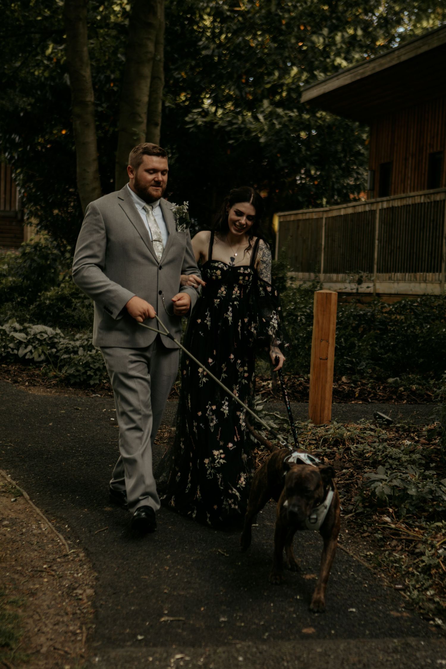 Couple in formal attire walking a dog on a leash along a garden path surrounded by lush greenery.