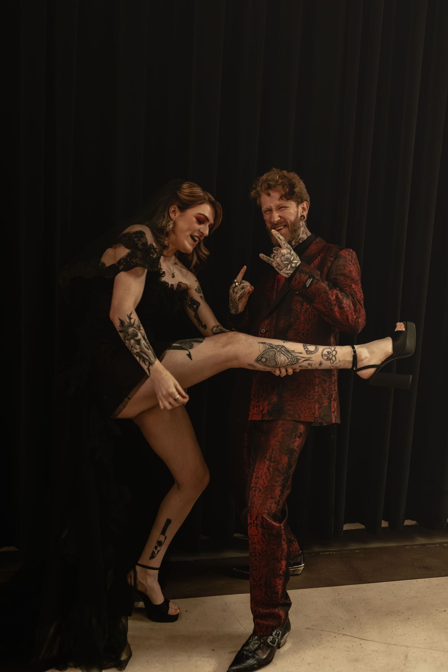 Tattooed couple in black and red wedding attire posing playfully against dark curtain backdrop.