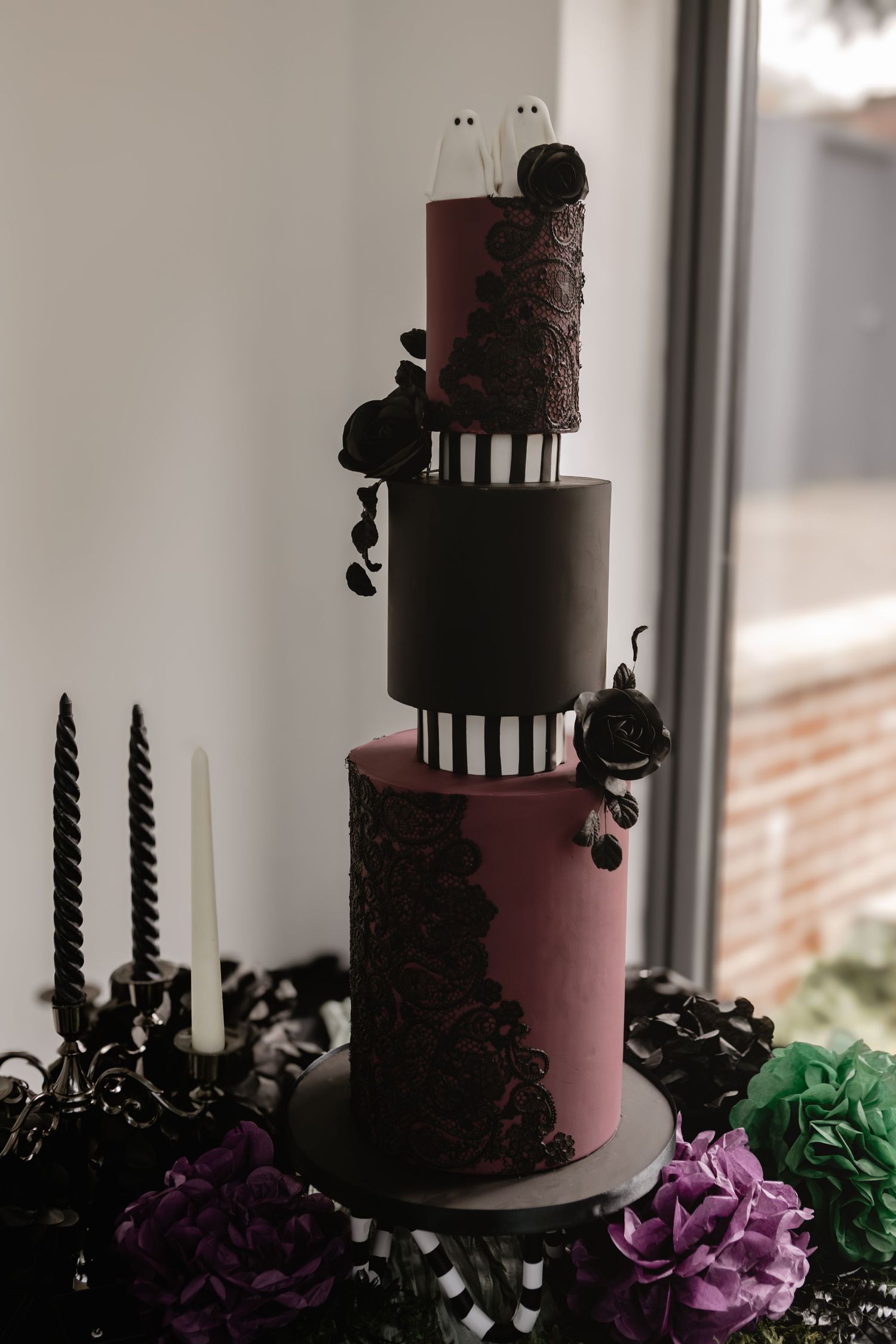 Four-tier gothic wedding cake in black and mauve with lace details, ghost toppers, and black rose decorations.