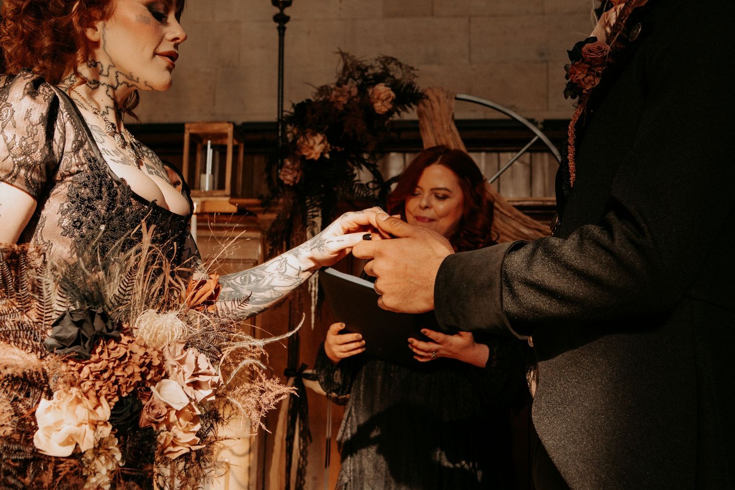 Officiant placing wedding ring on tattooed bride's finger during gothic wedding ceremony with floral arch.