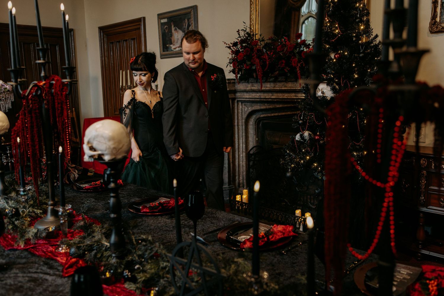 Couple in gothic wedding attire stand by candlelit altar with skulls, red garlands, and dark Christmas decor.