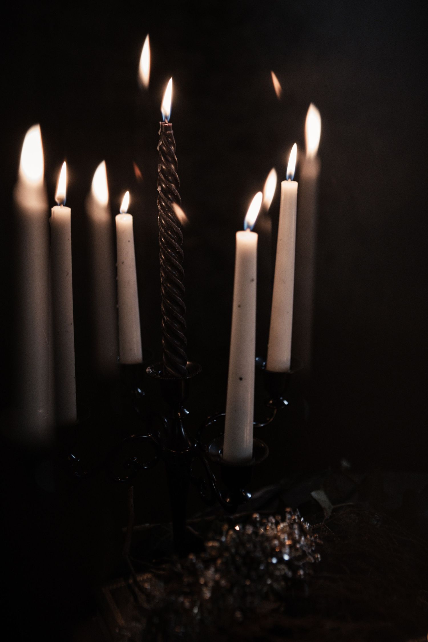 Dark candelabra with multiple lit white taper candles and one twisted black candle glowing against a dark background.