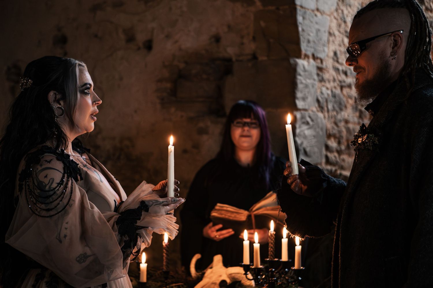 Gothic couple holding candles during dark wedding ceremony officiated by woman reading from ancient book.