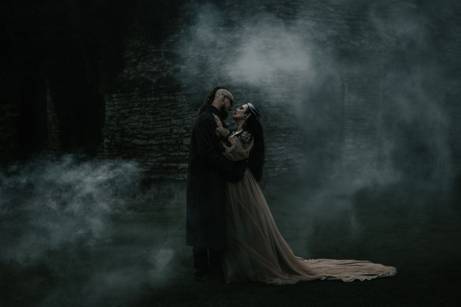 Gothic couple embracing in misty dark stone ruins, wearing dramatic black and flowing gown attire.