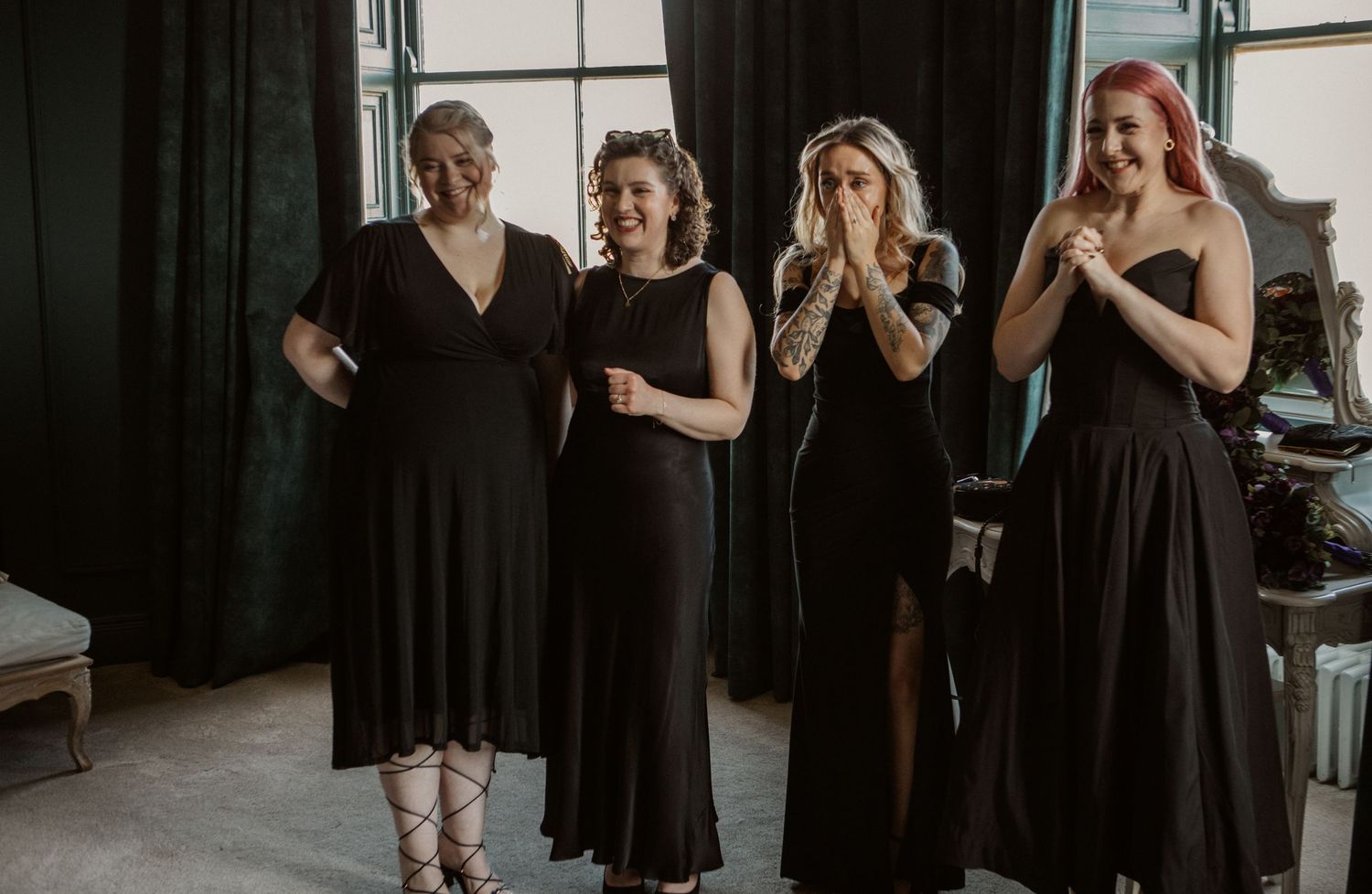 Four women in elegant black dresses stand together, smiling and reacting joyfully in a stylish indoor setting.