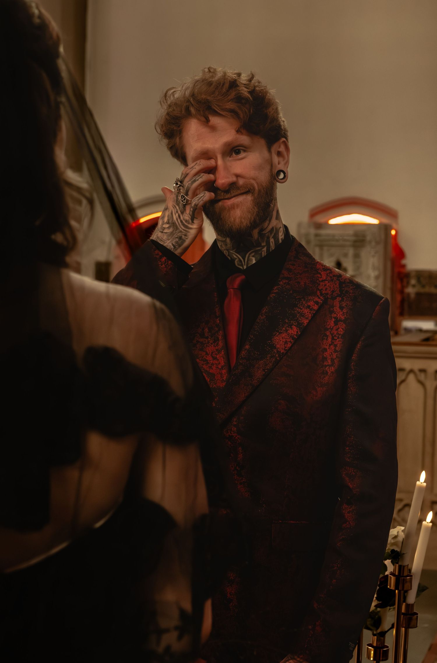 A bearded person with tattoos wearing a dark patterned jacket smiles and gestures in warm lighting.