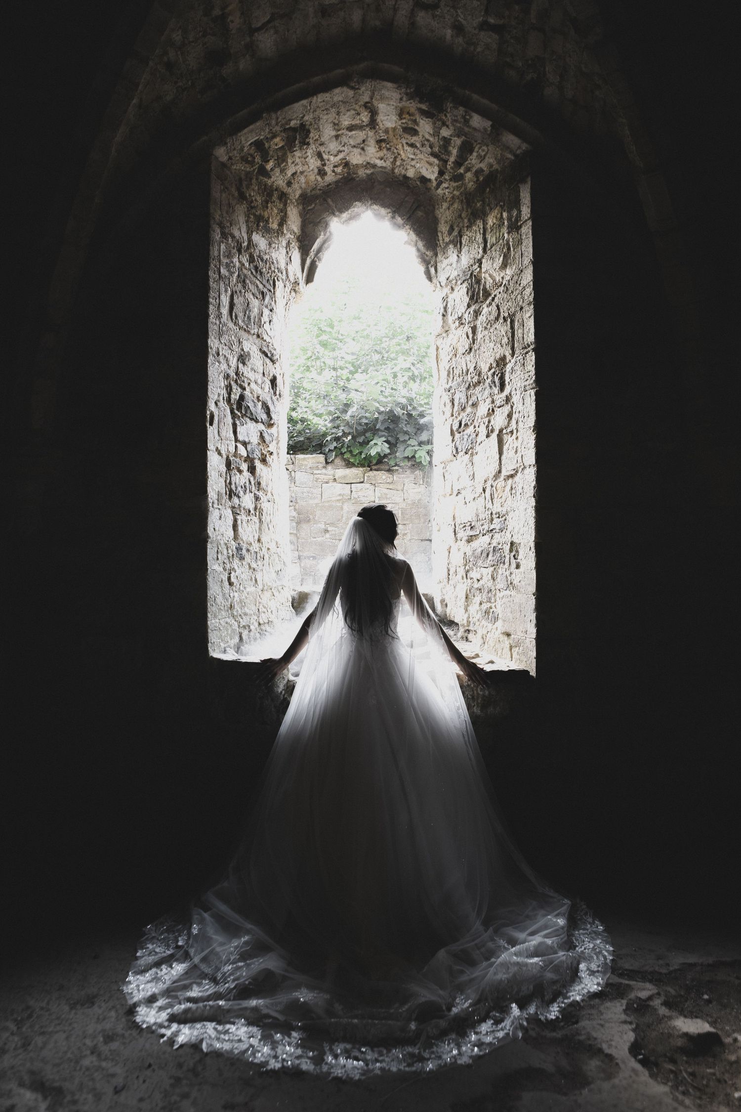 Bride in white ball gown standing silhouetted against arched stone window in ancient castle ruins.