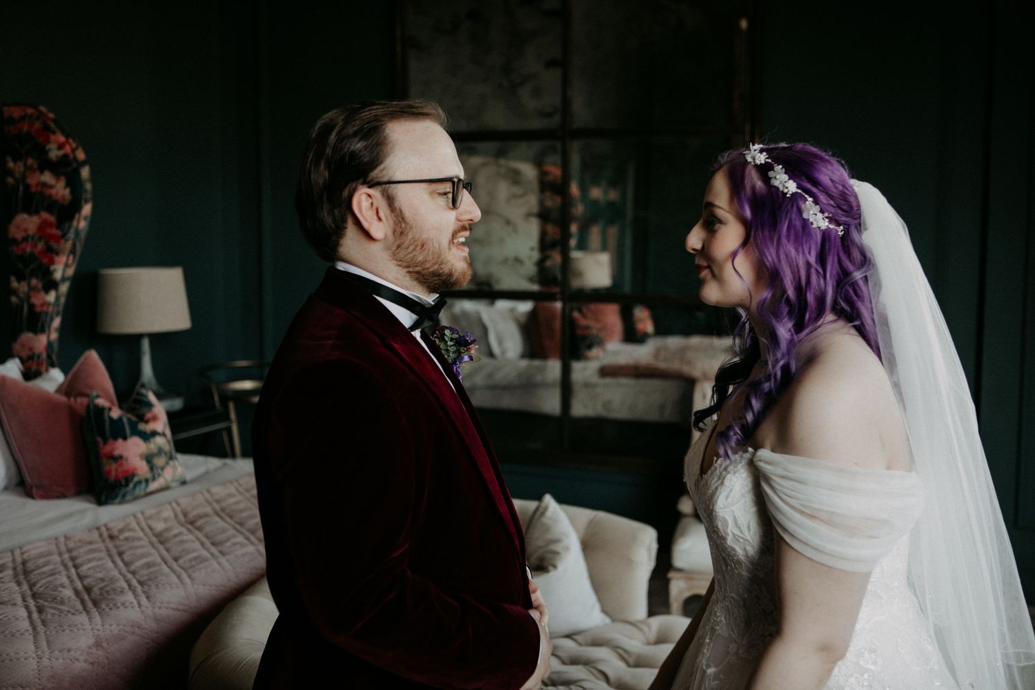 Bride with purple hair and groom in red velvet suit share a tender moment during their wedding ceremony indoors.