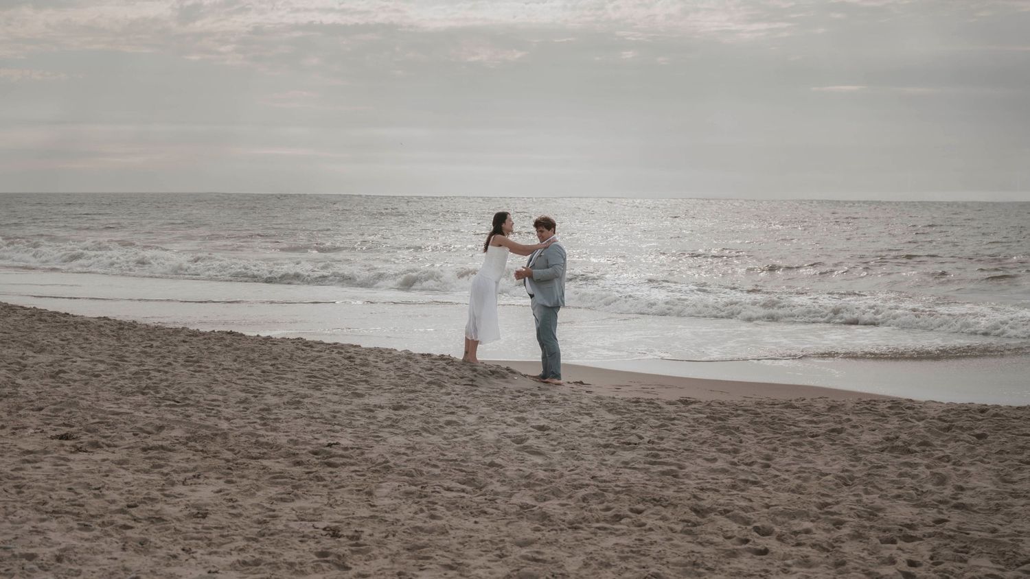 Couple in wedding attire standing together on empty beach with waves in background.