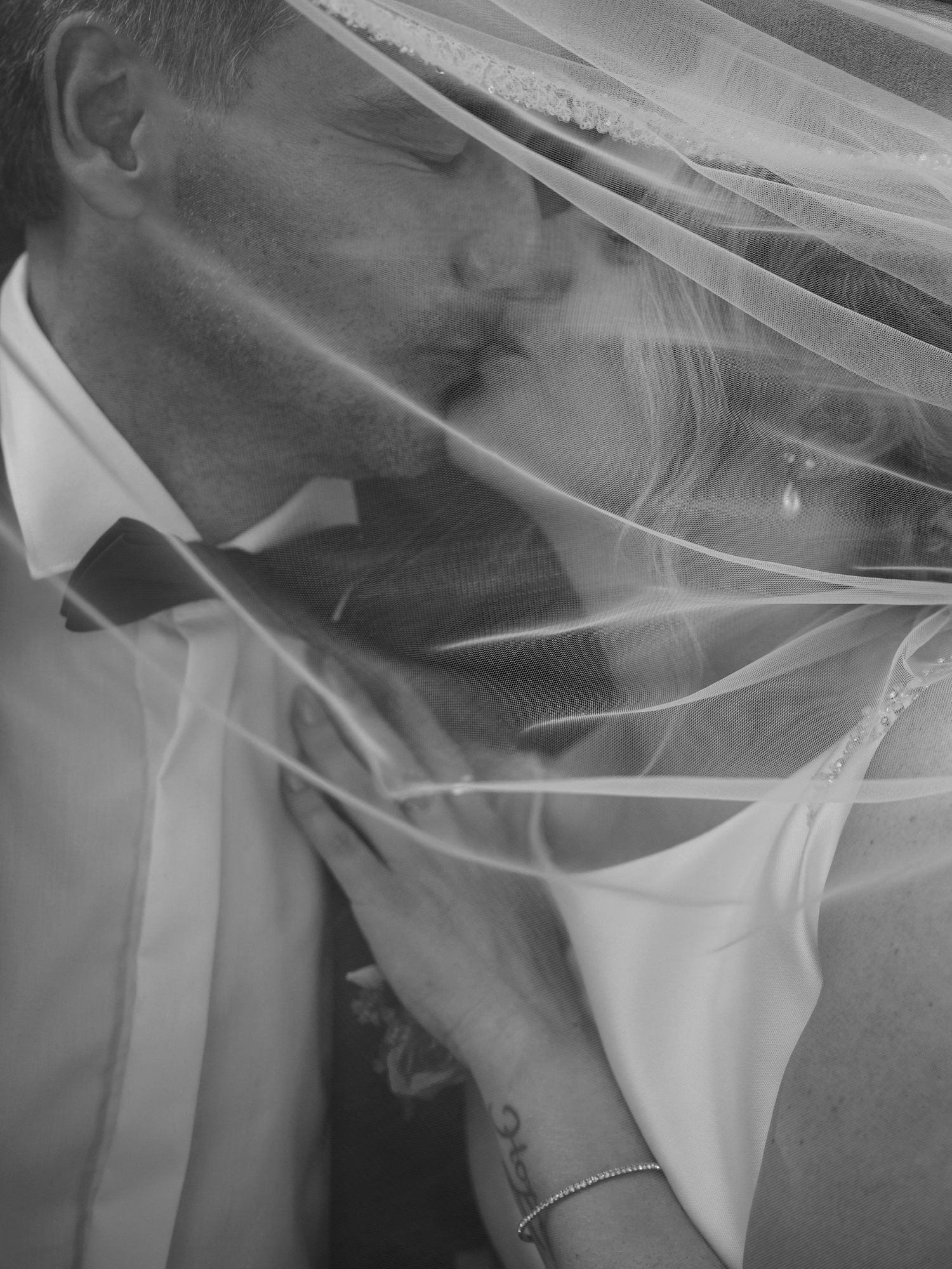 Black and white photo of bride and groom kissing under a flowing wedding veil, capturing a romantic intimate moment.