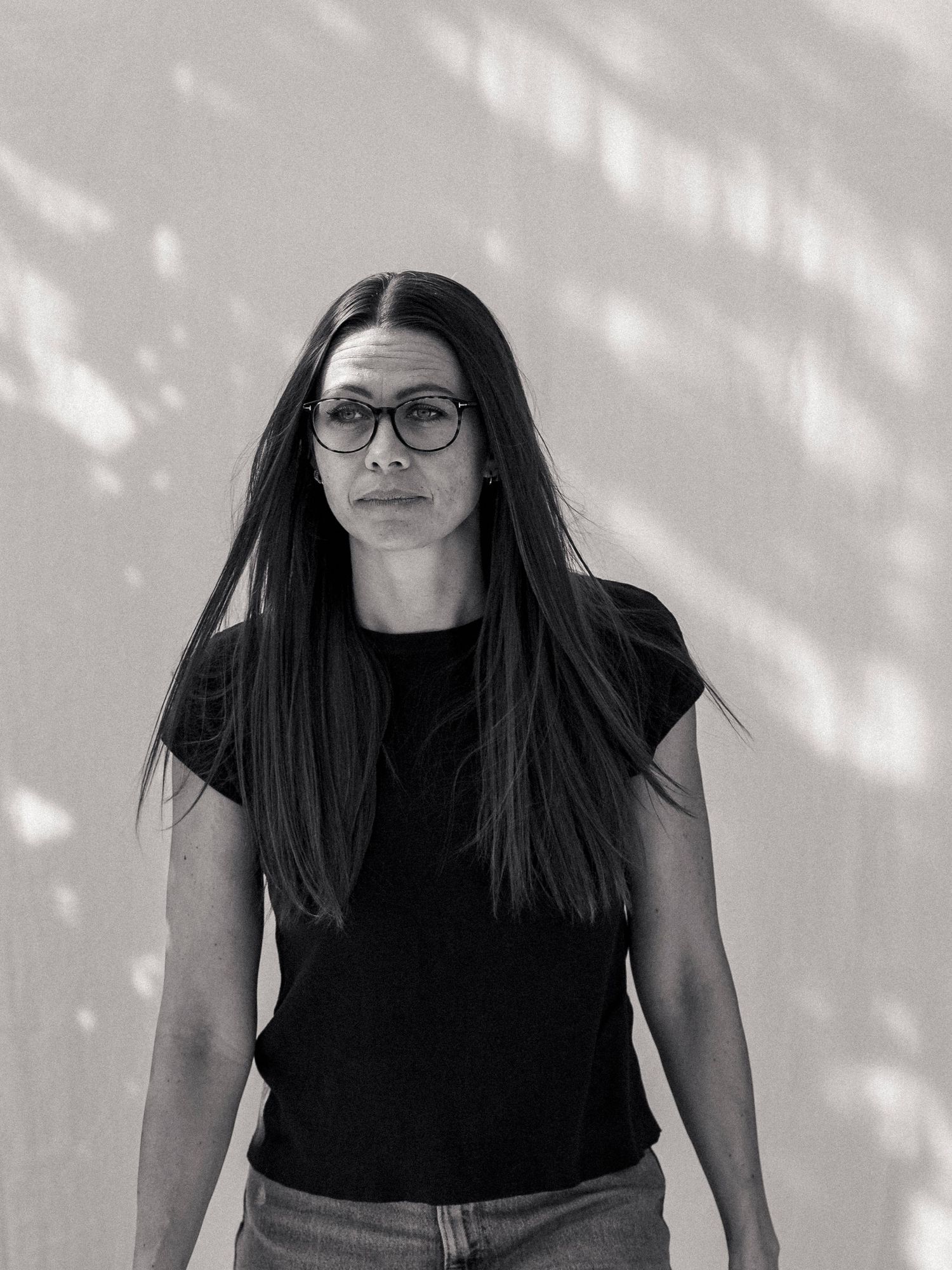 Black and white portrait of a woman with long dark hair and round glasses wearing a black t-shirt.