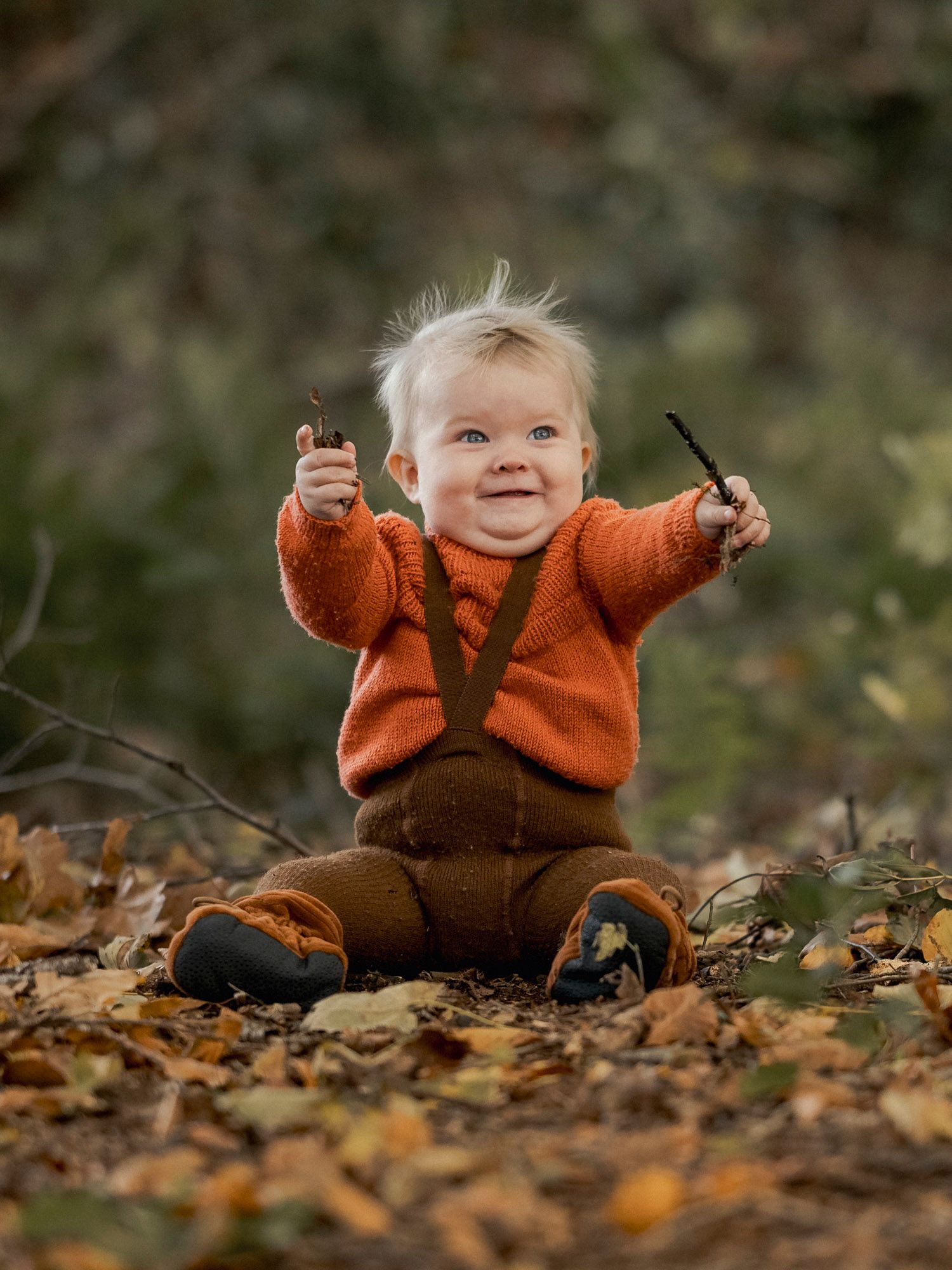 Happy toddler in orange sweater and brown suspenders sitting in autumn leaves holding sticks outdoors.