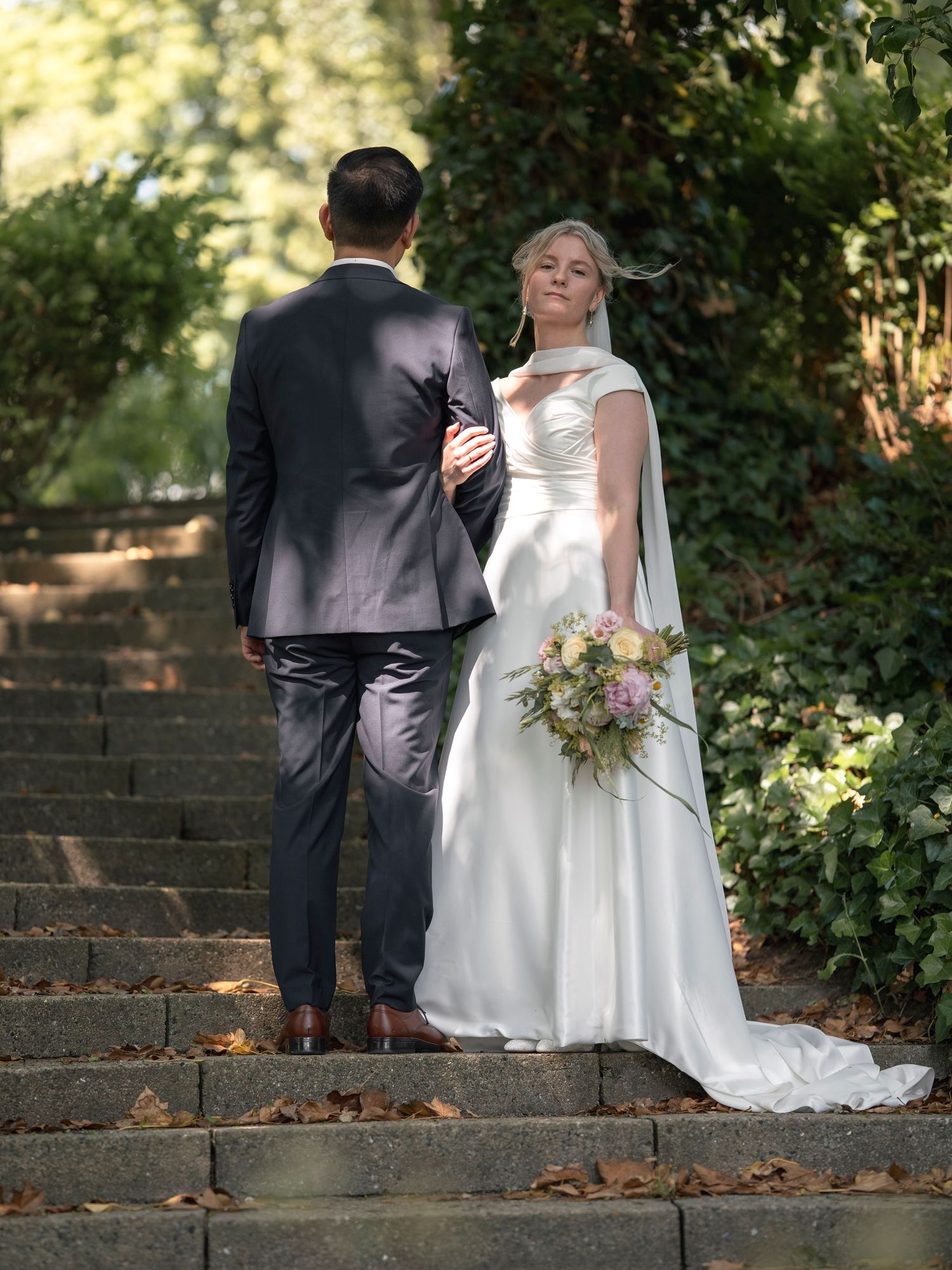 Bride in white gown holding bouquet stands with groom in gray suit on stone steps surrounded by lush greenery.