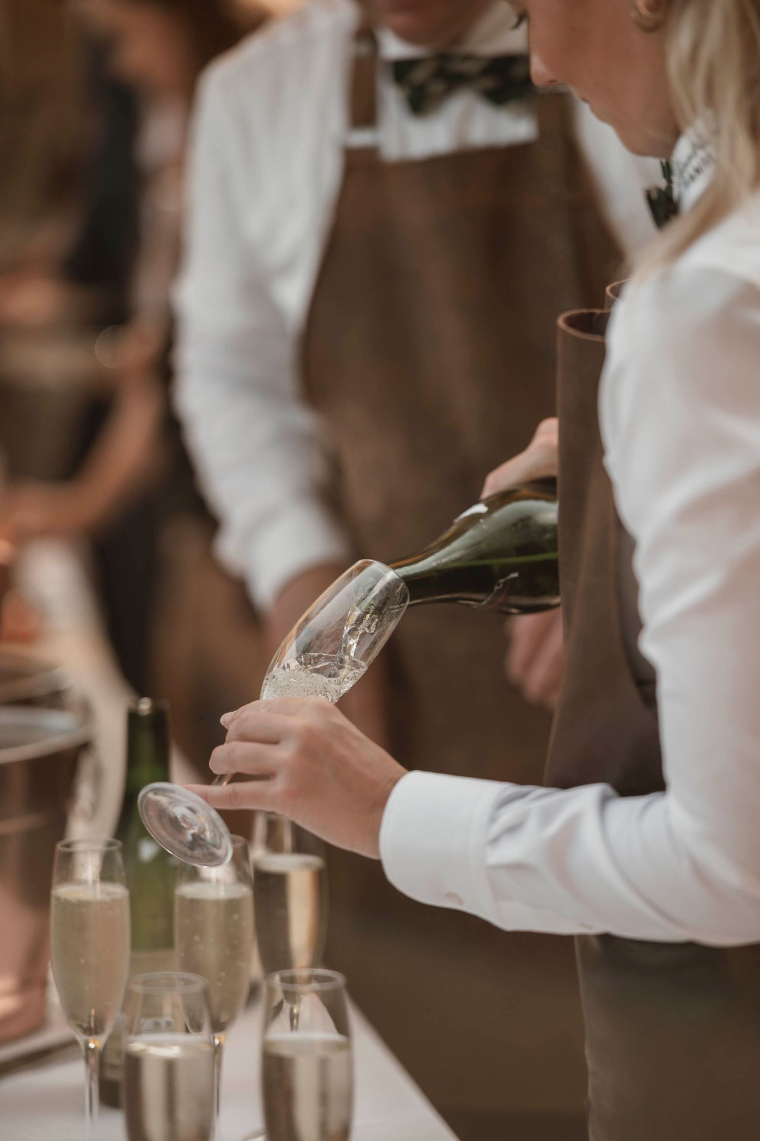 A bartender in a brown apron pours champagne into flutes at an elegant event.