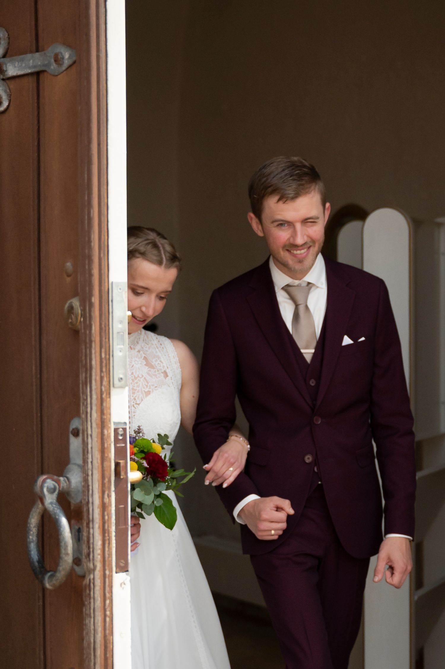 Smiling groom in burgundy suit escorts bride in white dress through wooden church doors on wedding day.
