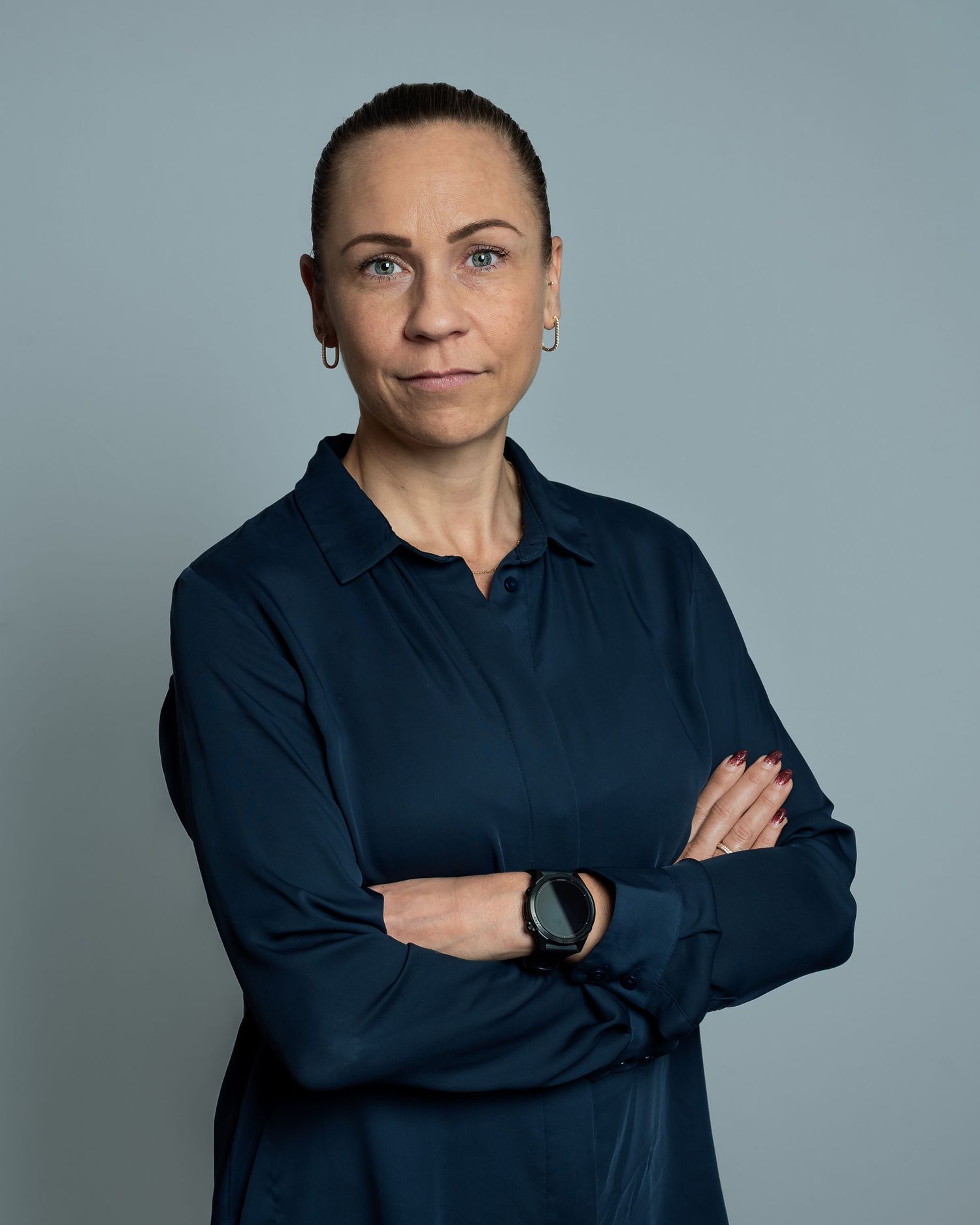 Professional woman in navy blue shirt with arms crossed, wearing a watch, against a gray background.