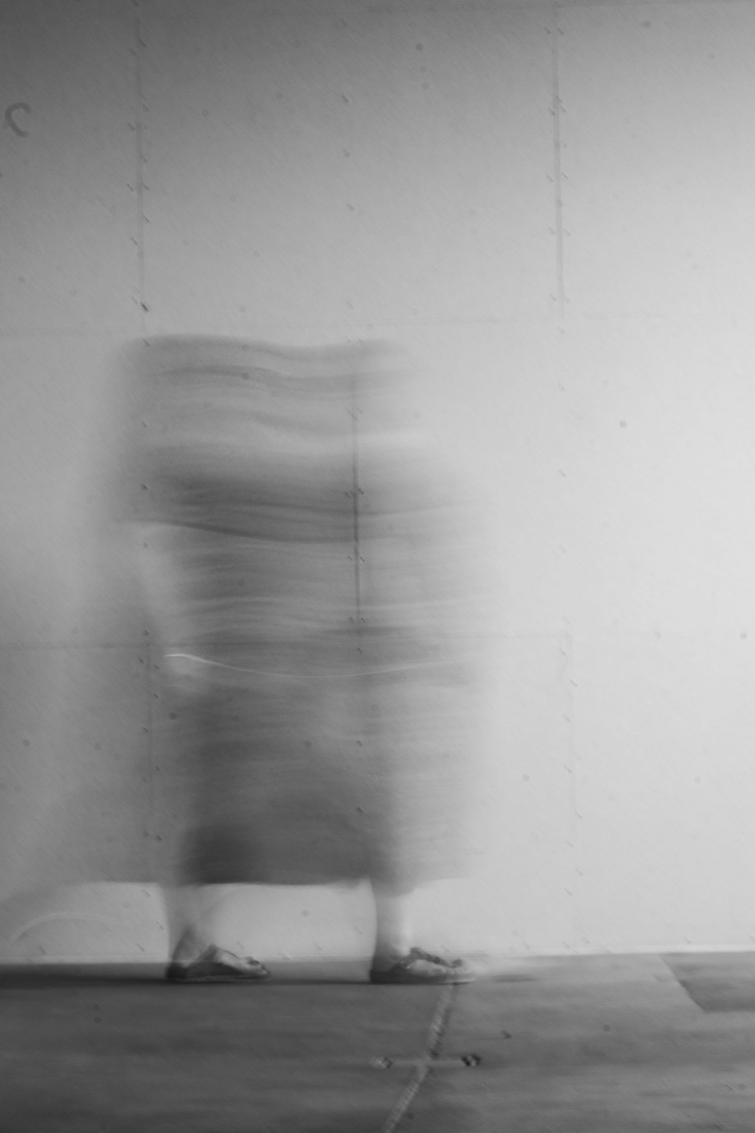 Blurred black and white long exposure photo of a person walking against a white wall.
