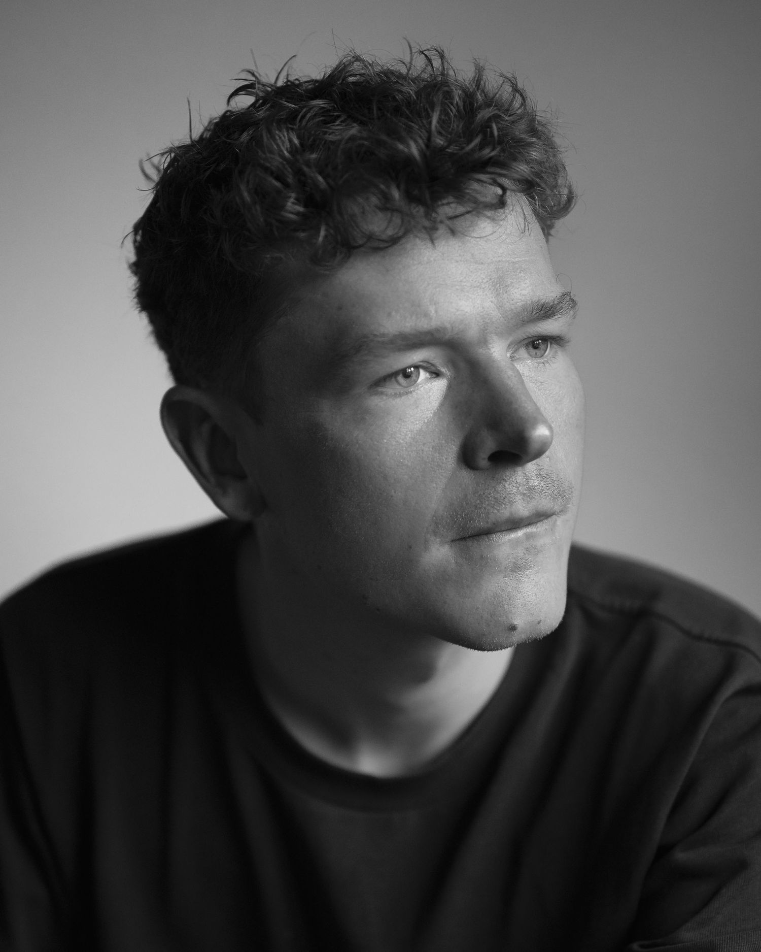 Black and white portrait of a young man with curly hair wearing a dark t-shirt, gazing thoughtfully to the side.