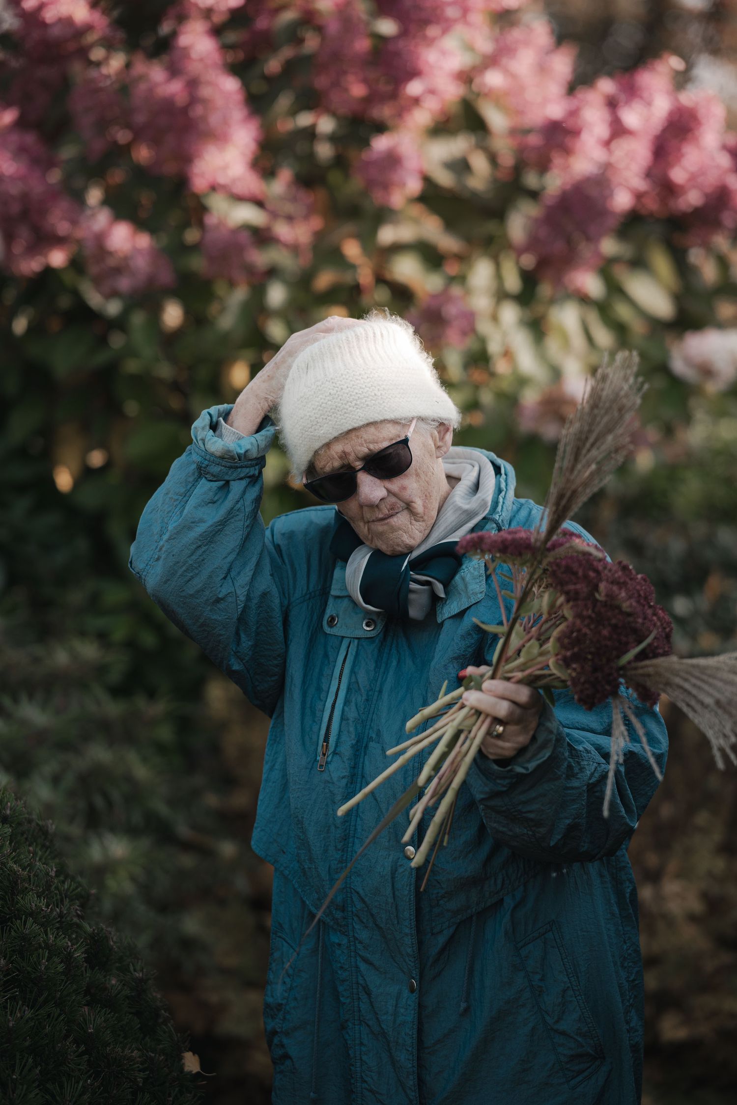 Elderly person in blue jacket and white hat holding dried flowers in a garden with pink blooms.