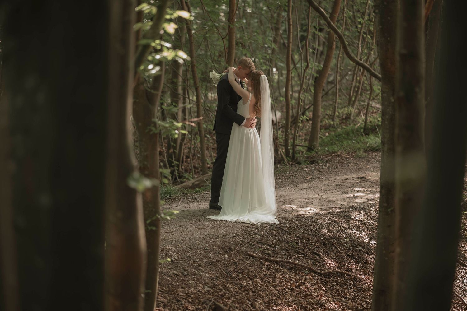 A bride and groom share a romantic kiss on a forest path, surrounded by tall trees and autumn leaves.