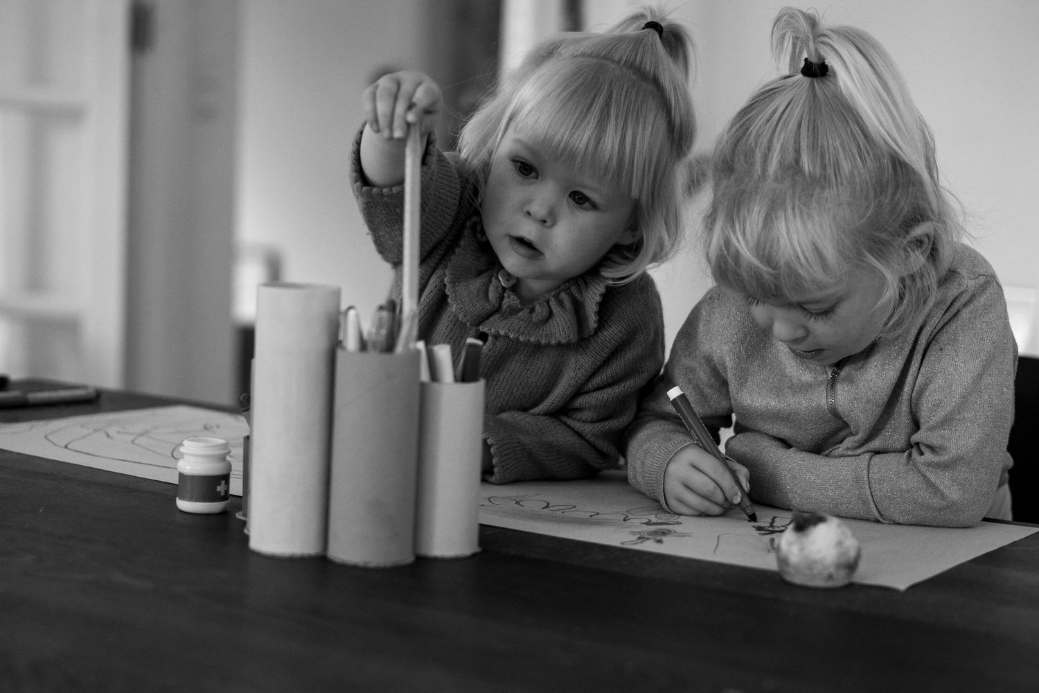 Two young blonde girls drawing and coloring together at a table with pencils and paint pots.