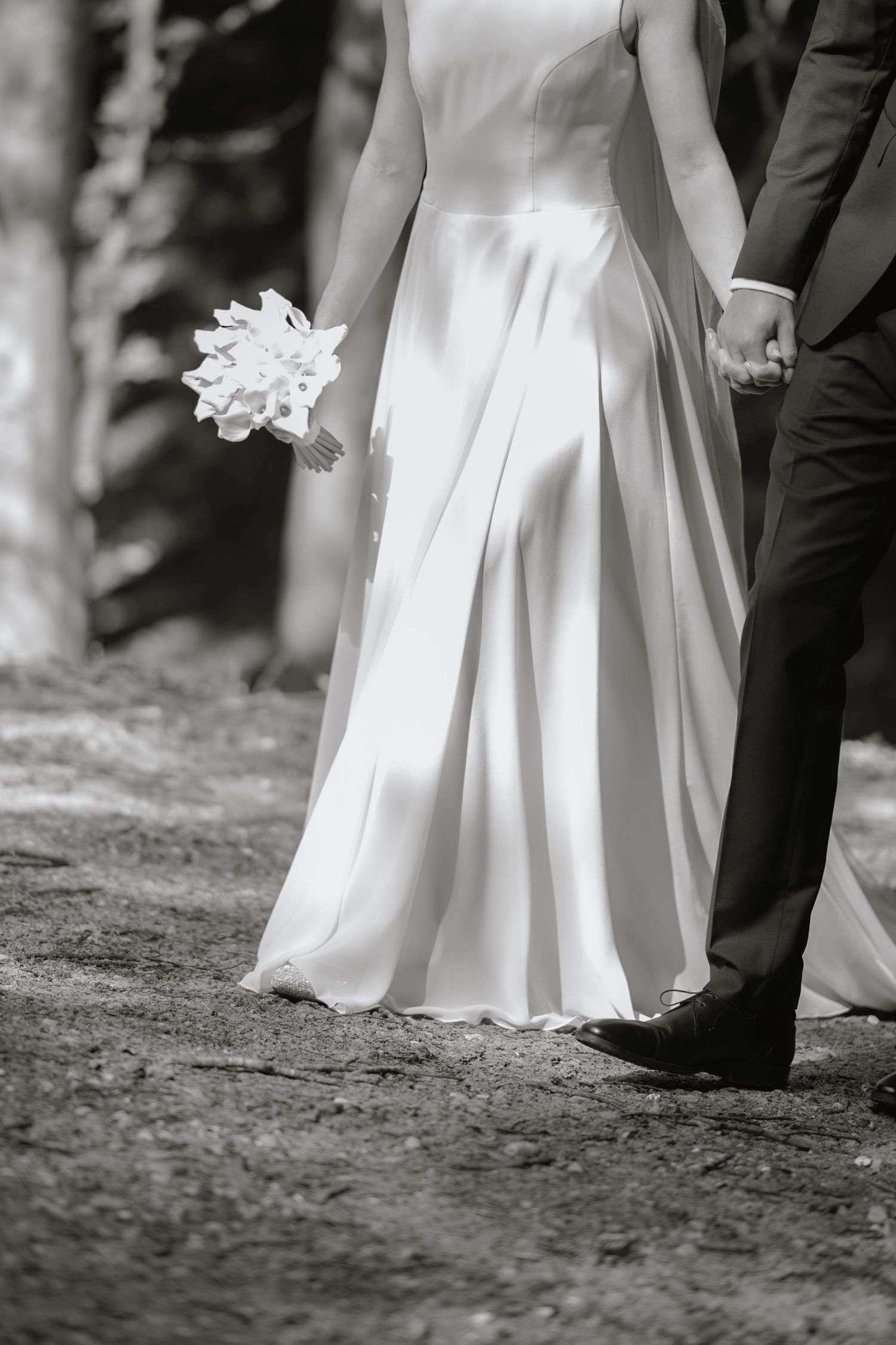 Black and white photo of bride in flowing white gown holding bouquet, walking hand in hand with groom in dark suit.